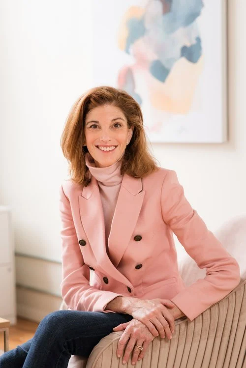 Woman wearing a pink blazer and turtleneck smiling while sitting on a beige chair in a room with a colorful abstract painting on the wall.