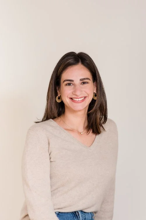 A smiling woman with shoulder-length brown hair, wearing a beige sweater, gold earrings, and a delicate necklace, standing against a plain light-colored background.