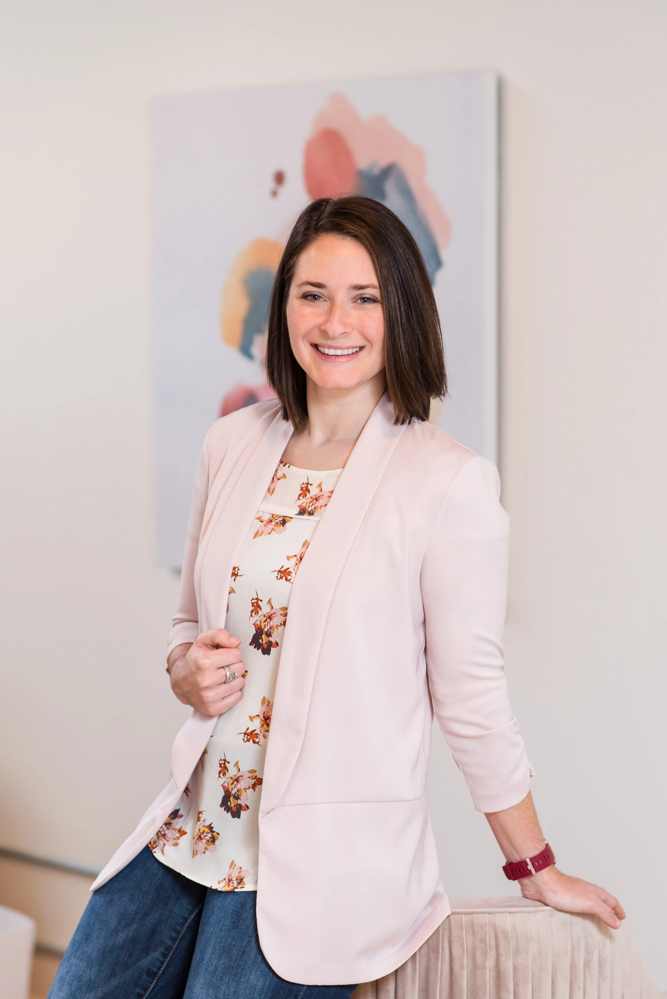 A woman with shoulder-length brown hair, wearing a cream blazer over a floral blouse, smiling and standing in an indoor setting with an abstract painting in the background. She is casually leaning on a piece of furniture and looking at the camera.