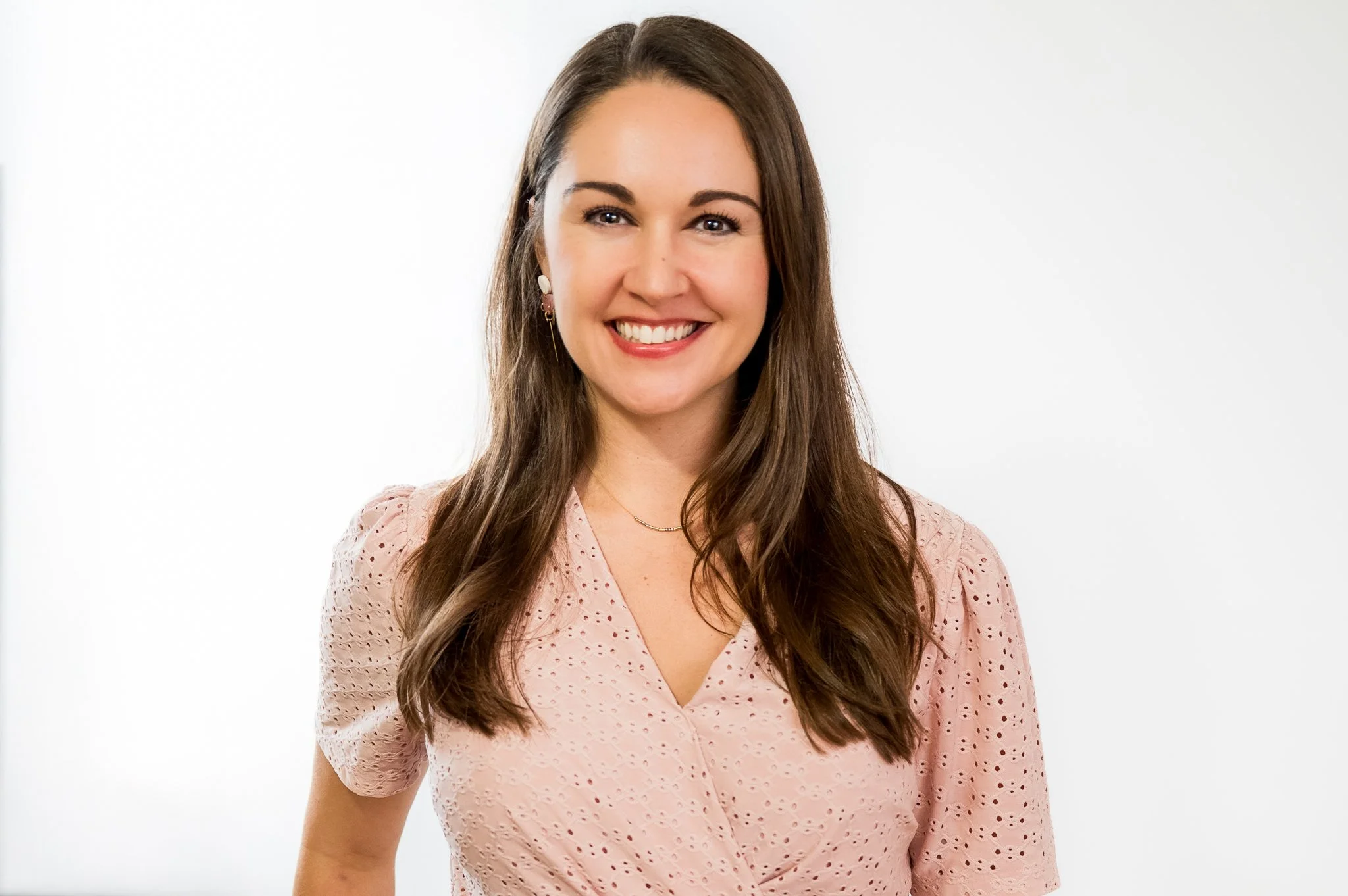 A smiling woman with long brown hair wearing a pink eyelet dress stands in front of a plain white background.