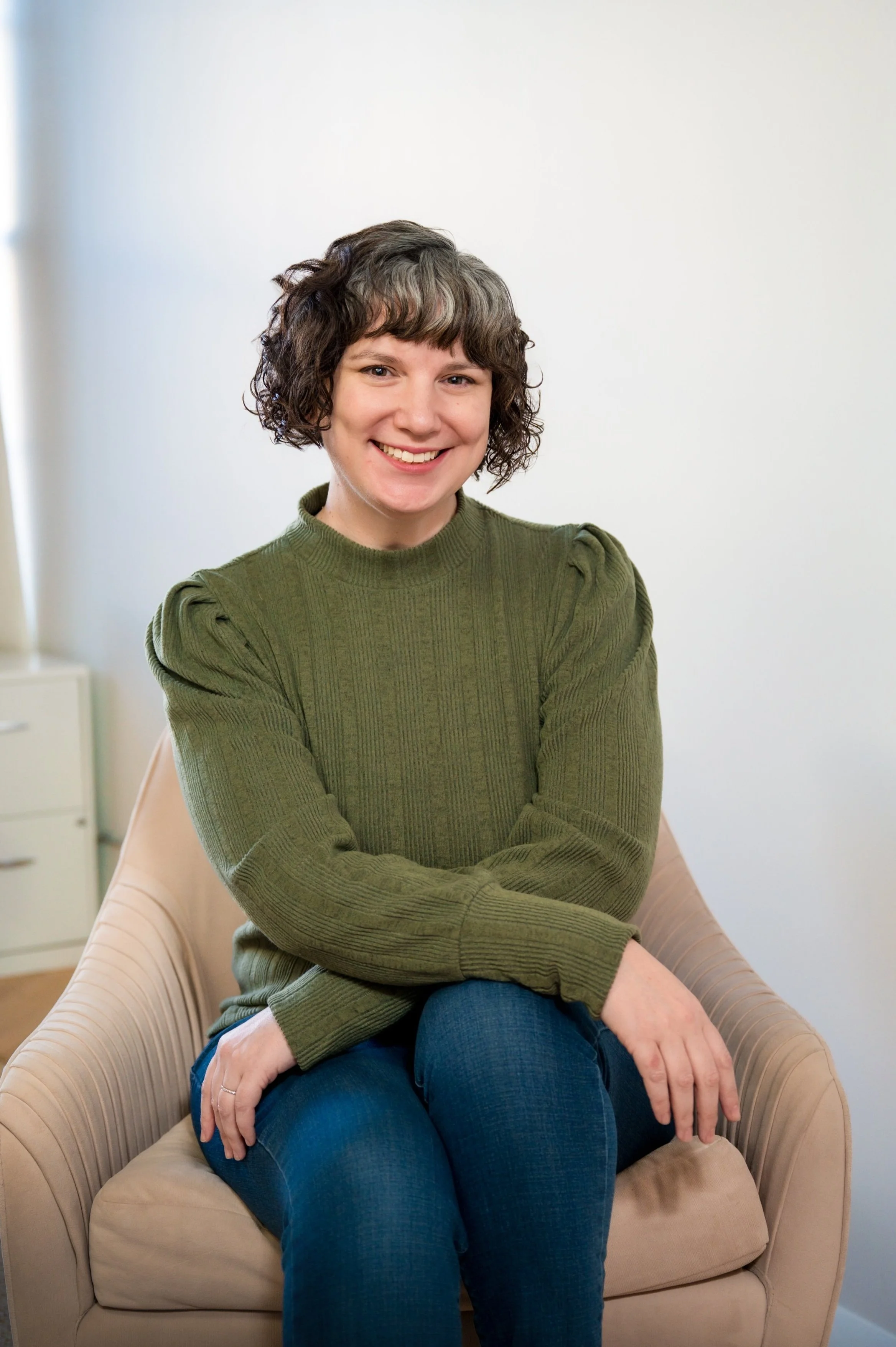 A woman with short, curly dark hair and a bright smile, sitting on a beige armchair with crossed legs, wearing a green sweater and blue jeans, in a room with plain white walls and a small white dresser in the background.