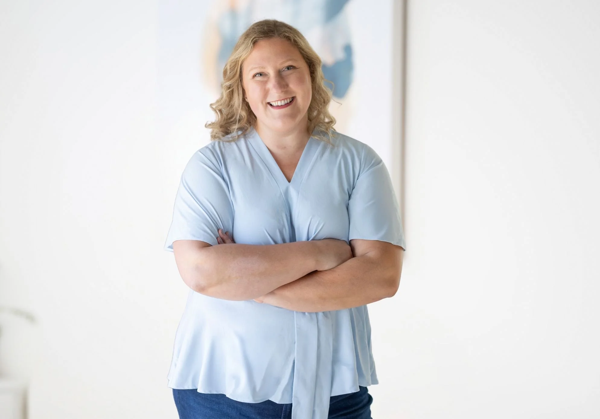 A woman with blond curly hair smiling and crossing her arms, wearing a light blue medical scrubs and standing in front of a white background with a blurred blue and white painting.