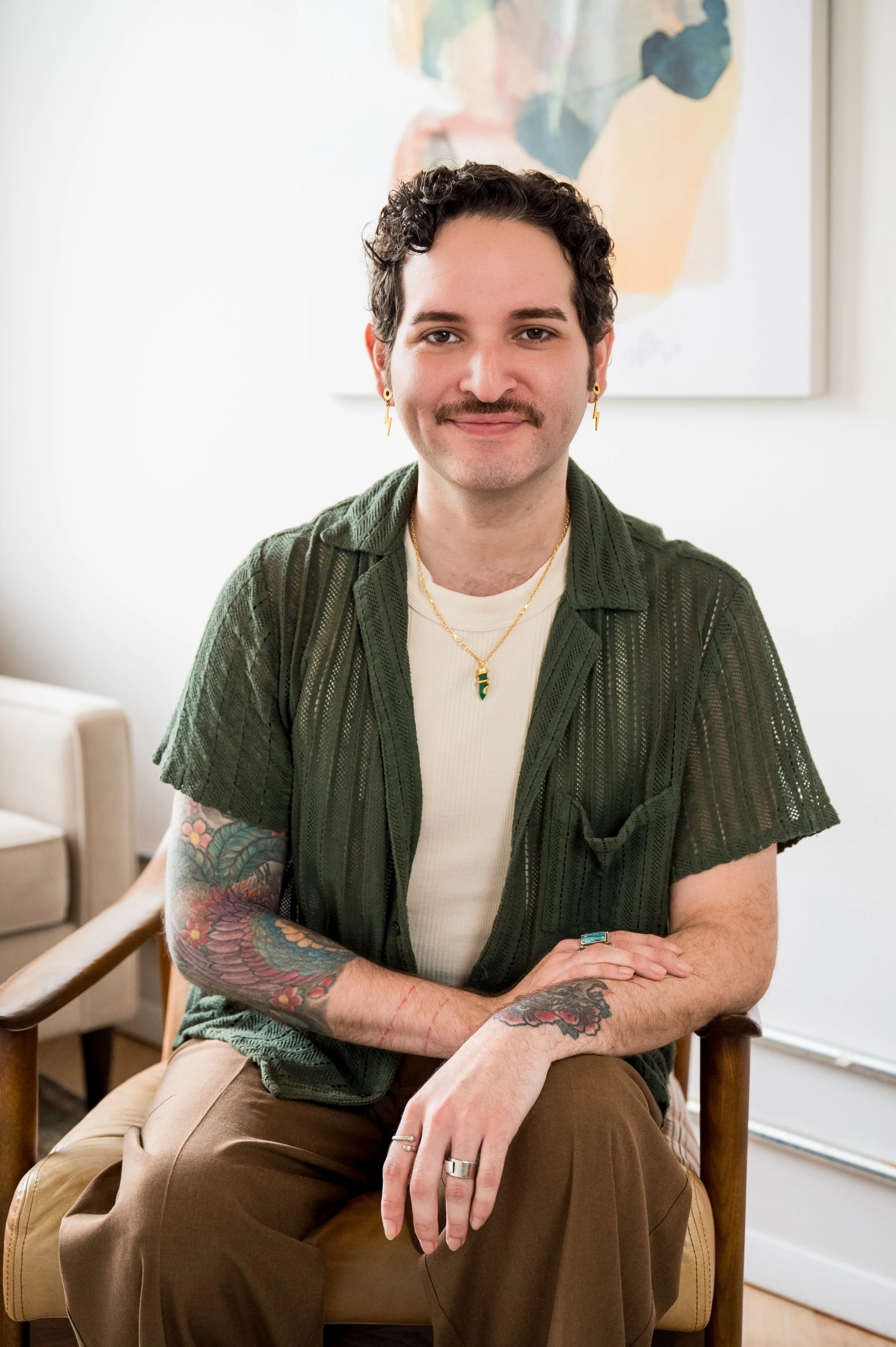 A young man with curly dark hair, mustache, and earrings is sitting on a wooden chair with a light-colored cushion in a bright room. He is wearing a white shirt and an open green cardigan, showing colorful tattoos on his arms. His hands are resting on his lap, adorned with rings. In the background, a colorful abstract painting is visible on a white wall.