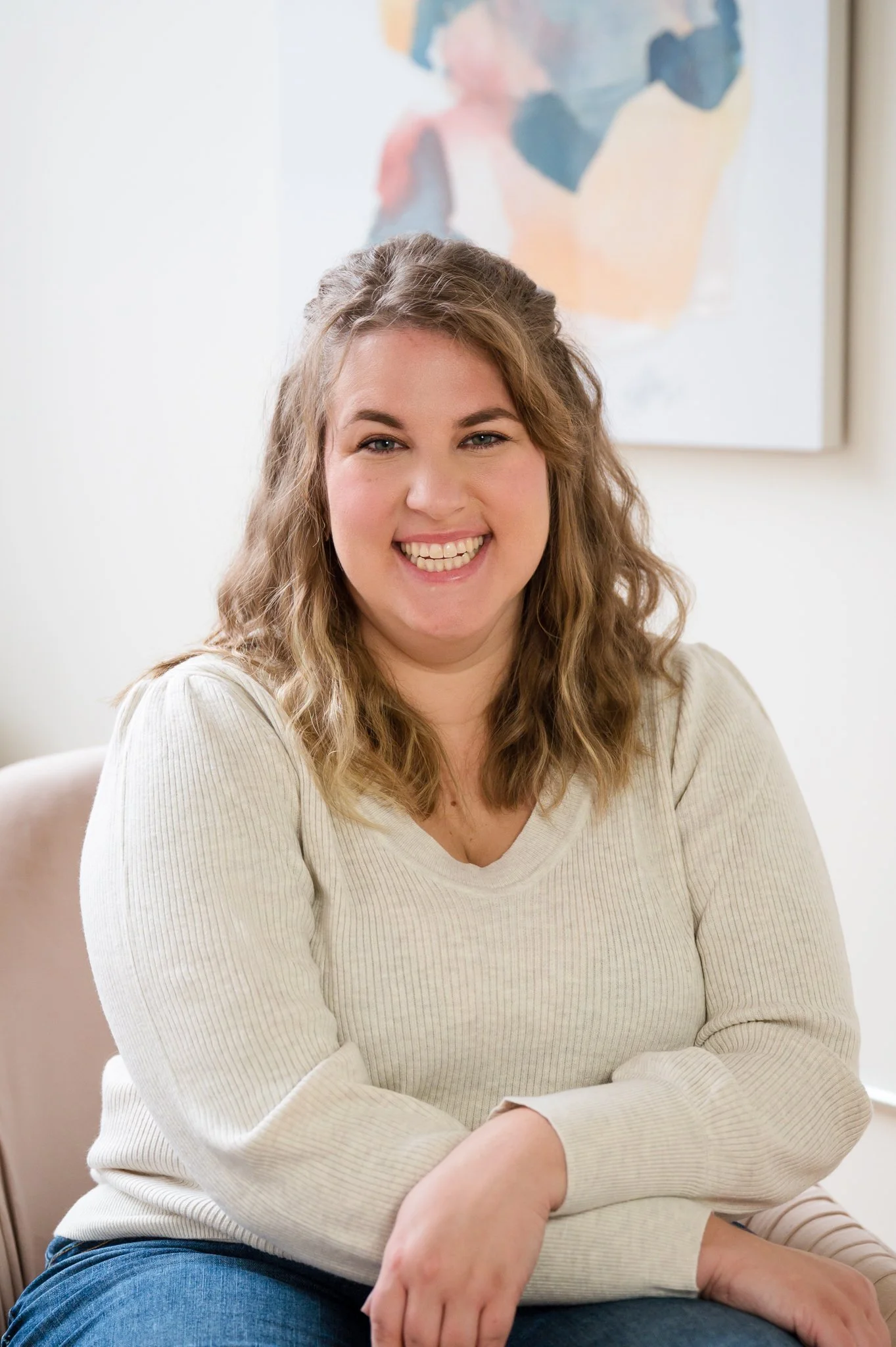 A woman sitting on a sofa in a cream-colored sweater, smiling at the camera with a colorful abstract painting behind her.