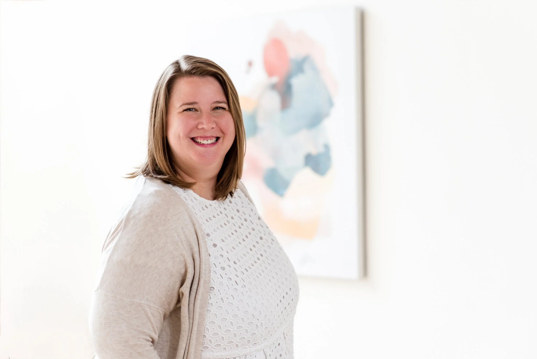 A smiling woman with shoulder-length brown hair standing in front of a colorful abstract painting on a white wall.