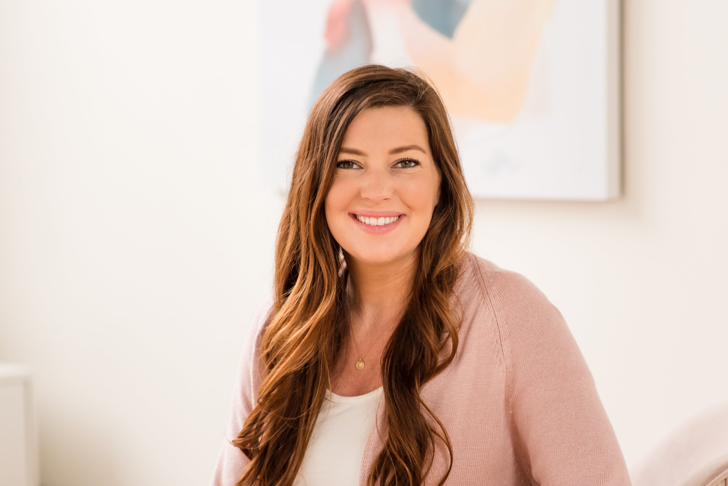 A smiling woman with long wavy hair wearing a light pink cardigan and a white top, sitting in a well-lit room with a framed abstract art piece on the wall behind her.