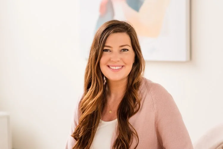 A woman with long brown hair and a pink cardigan smiling at the camera, sitting in a bright room with a painting on the wall behind her.