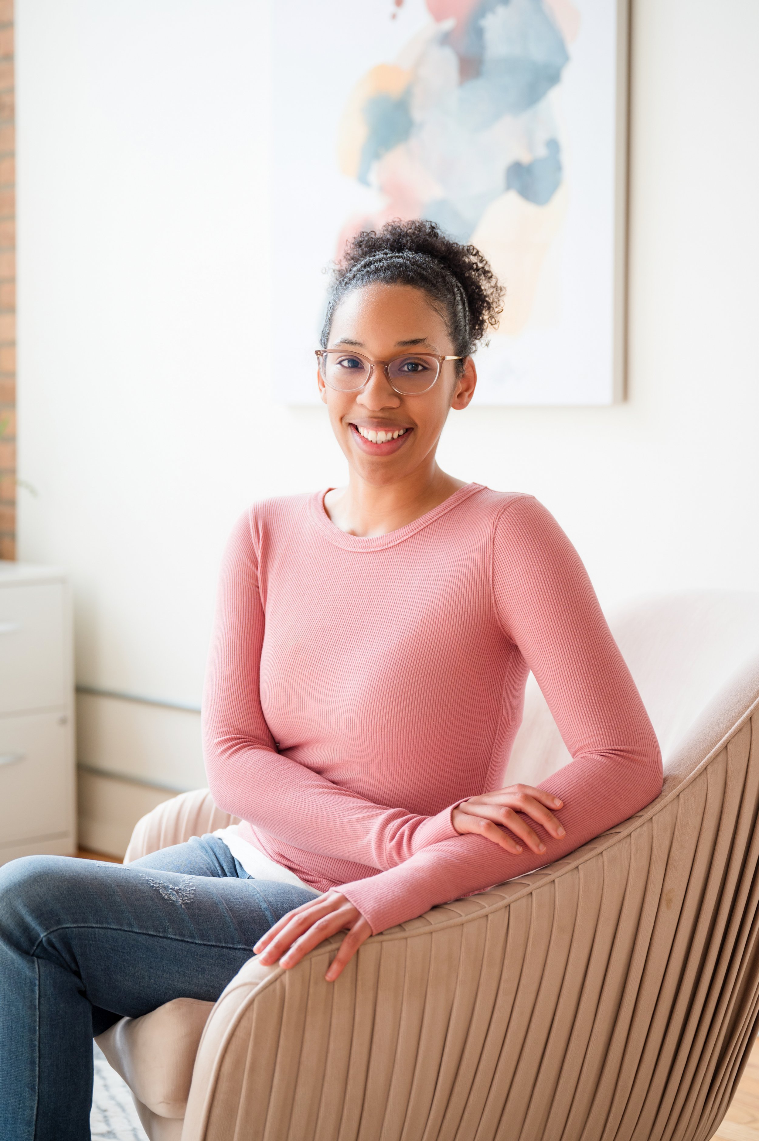 A woman with glasses and curly hair sitting on a beige chair in a room with a colorful abstract painting on the wall behind her, smiling at the camera.