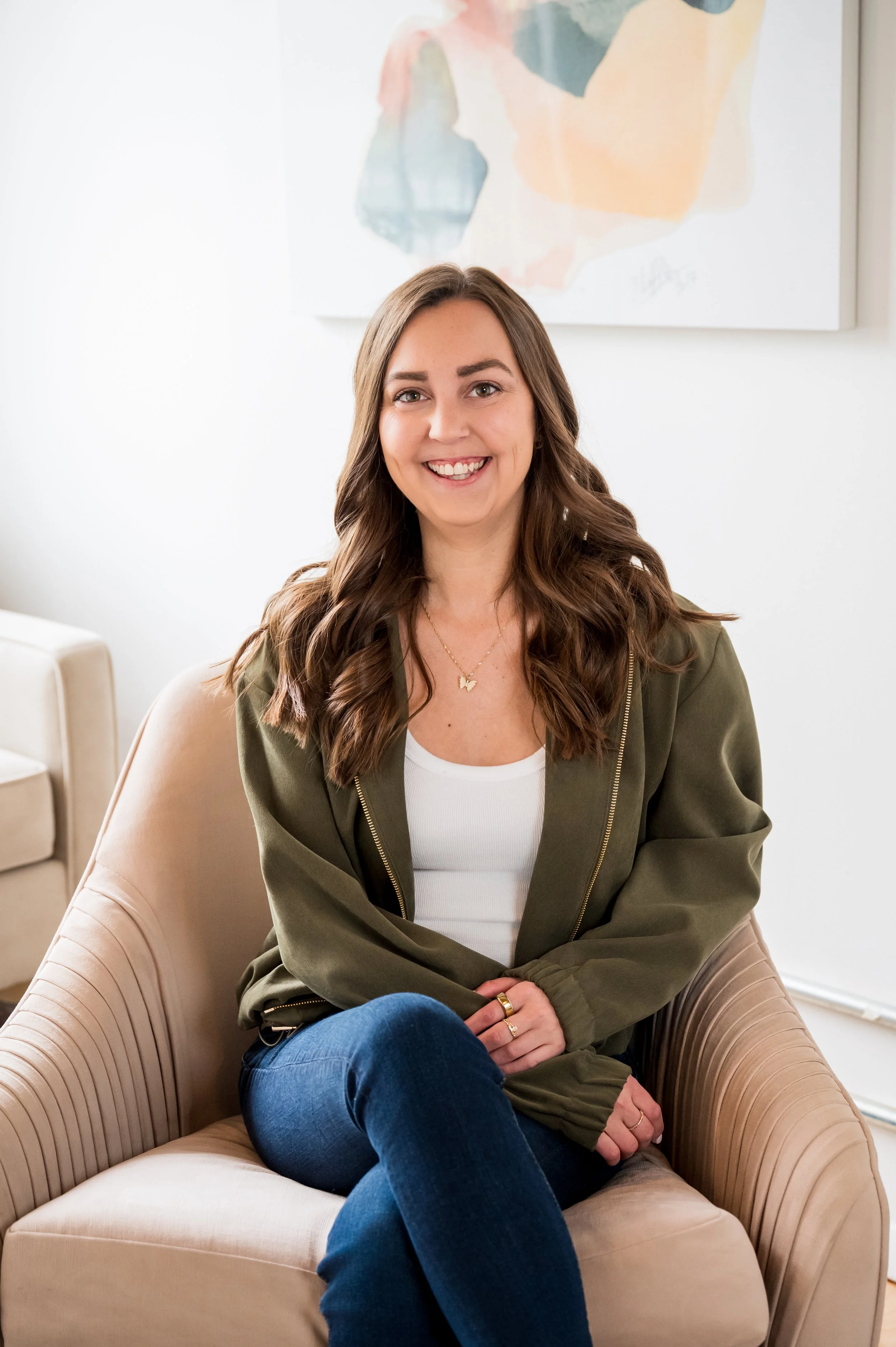A young woman with long, wavy brown hair and light skin sitting on a beige armchair, smiling at the camera. She is wearing a white top, an olive green zip-up jacket, and blue jeans. Behind her, there is a white wall with an abstract colorful painting.