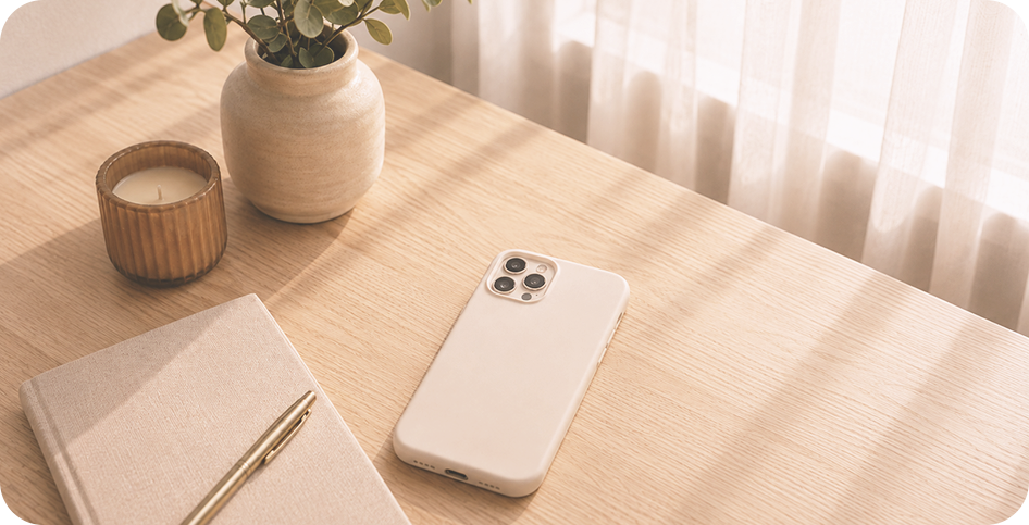 A wooden desk with a potted plant, a candle, a closed notebook with a gold pen, and a white smartphone, with sheer curtains in the background.