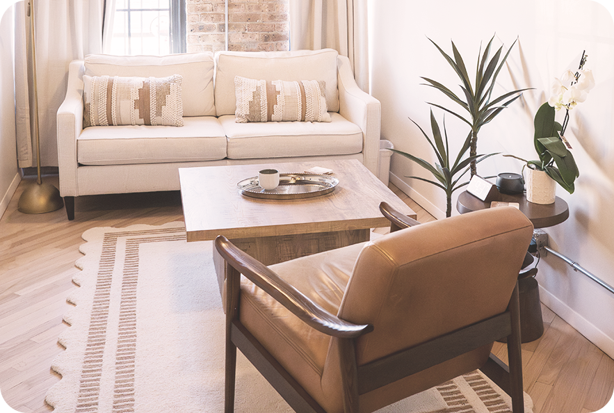 Cozy living room with cream sofa, wooden coffee table, tan armchair, potted plants, and natural light.