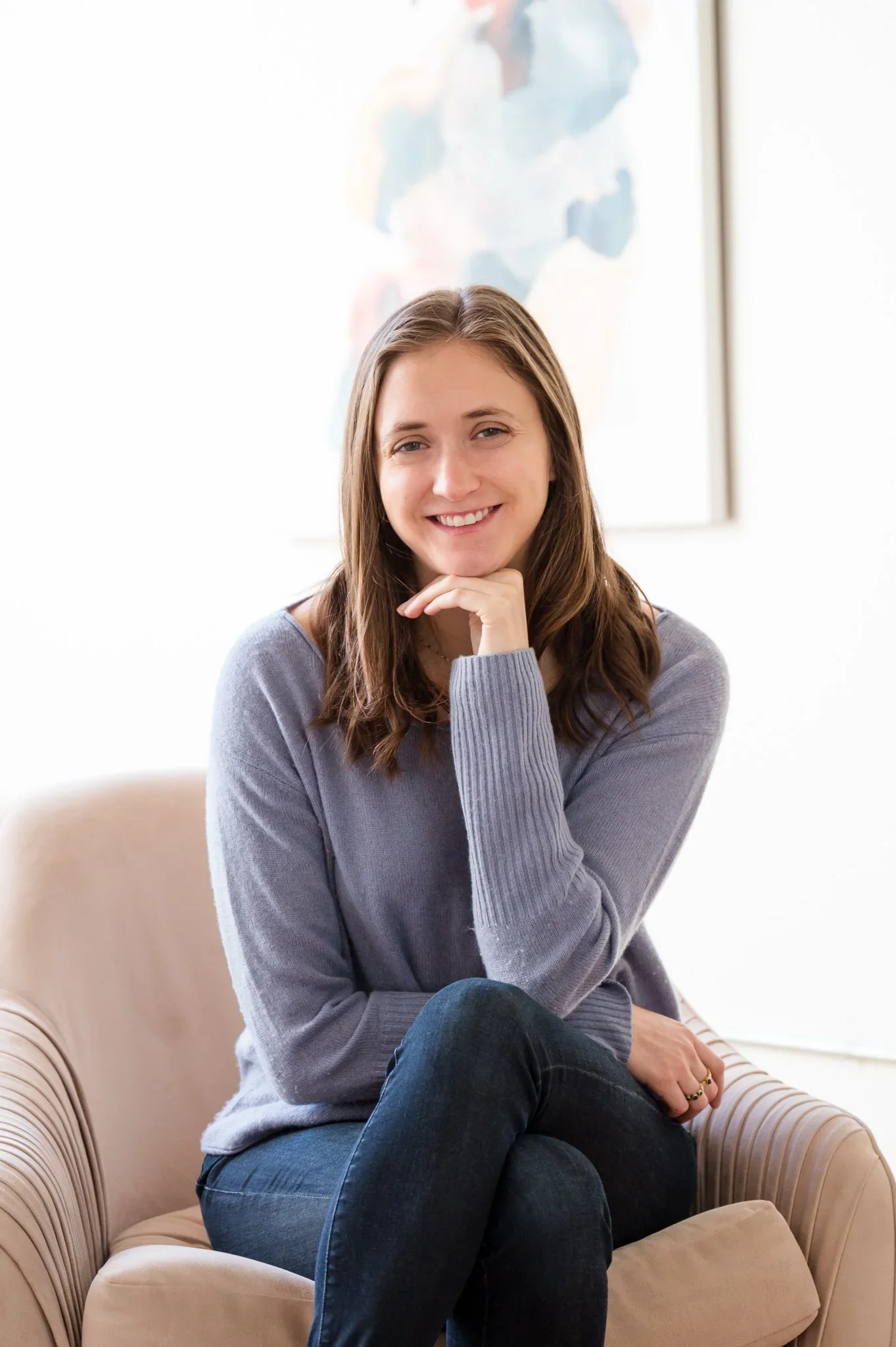 A young woman with long brown hair smiling, sitting on a beige armchair, wearing a gray sweater and dark jeans, with her hand resting under her chin. In the background, there is a light blue abstract painting.