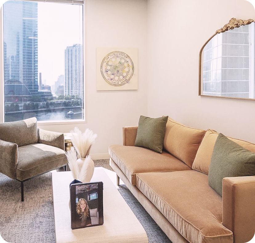 Living room with beige sofa, green pillows, gray armchair, white coffee table, large window showing cityscape, decorative mirror, abstract wall art, and a framed photo on the table.