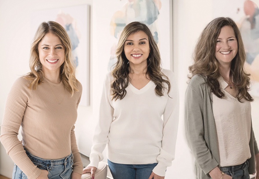 Three women standing and smiling in a well-lit room with abstract artwork on the wall behind them.