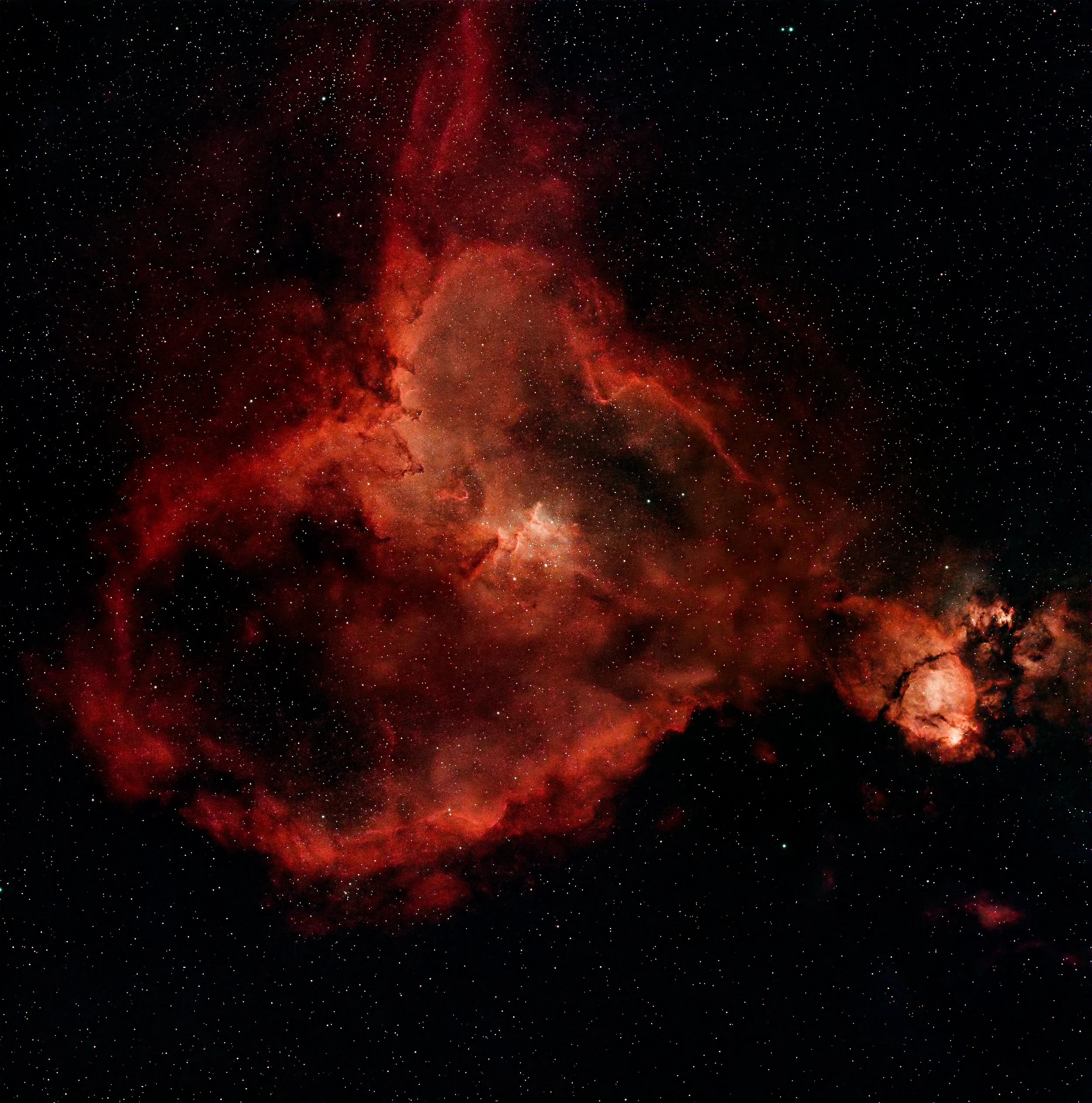 A view of outer space featuring numerous small white stars scattered across a dark background with a large, bright, reddish nebula in the center and surrounding gaseous clouds.