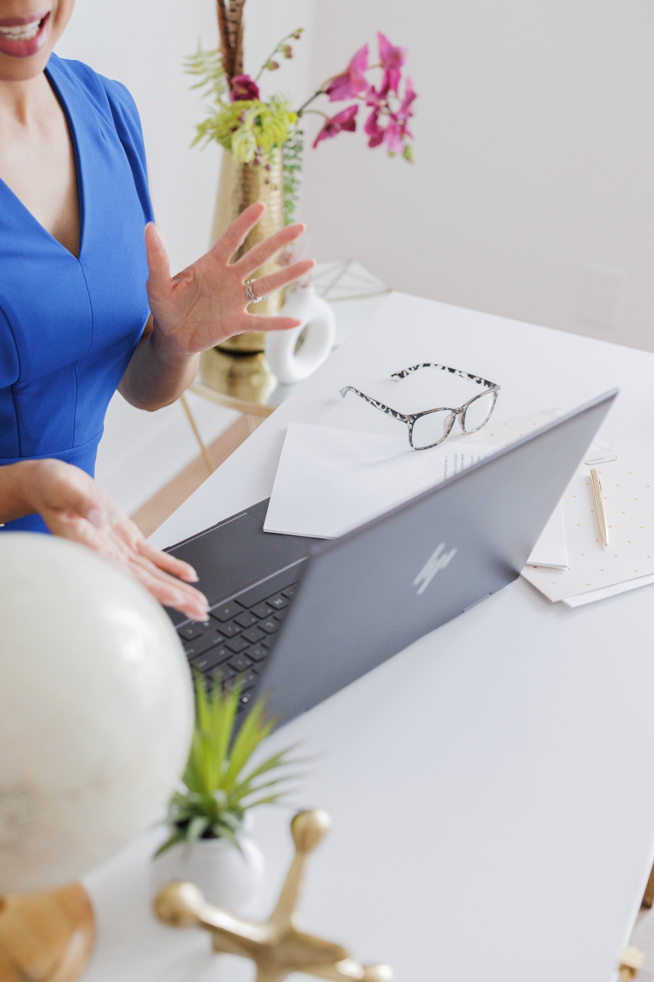 Partially visible woman in a blue top gesturing with her hand near a white desk. On the desk are a pair of eyeglasses, a paper with printed text, a gold-colored pen, and a laptop. In the foreground, blurred vase with white and gold decorative elements and a green plant. In the background, a vase with pink and green flowers on a side table.