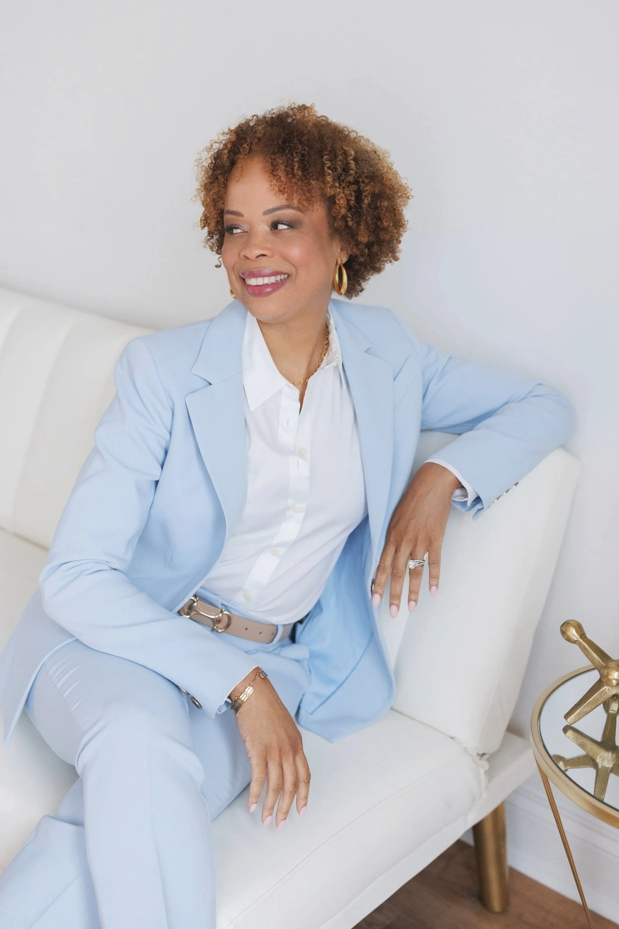 A woman with curly brown hair, wearing a light blue blazer and pants, and a white shirt, sitting on a white sofa with a gold side table beside her, smiling and looking to the side.