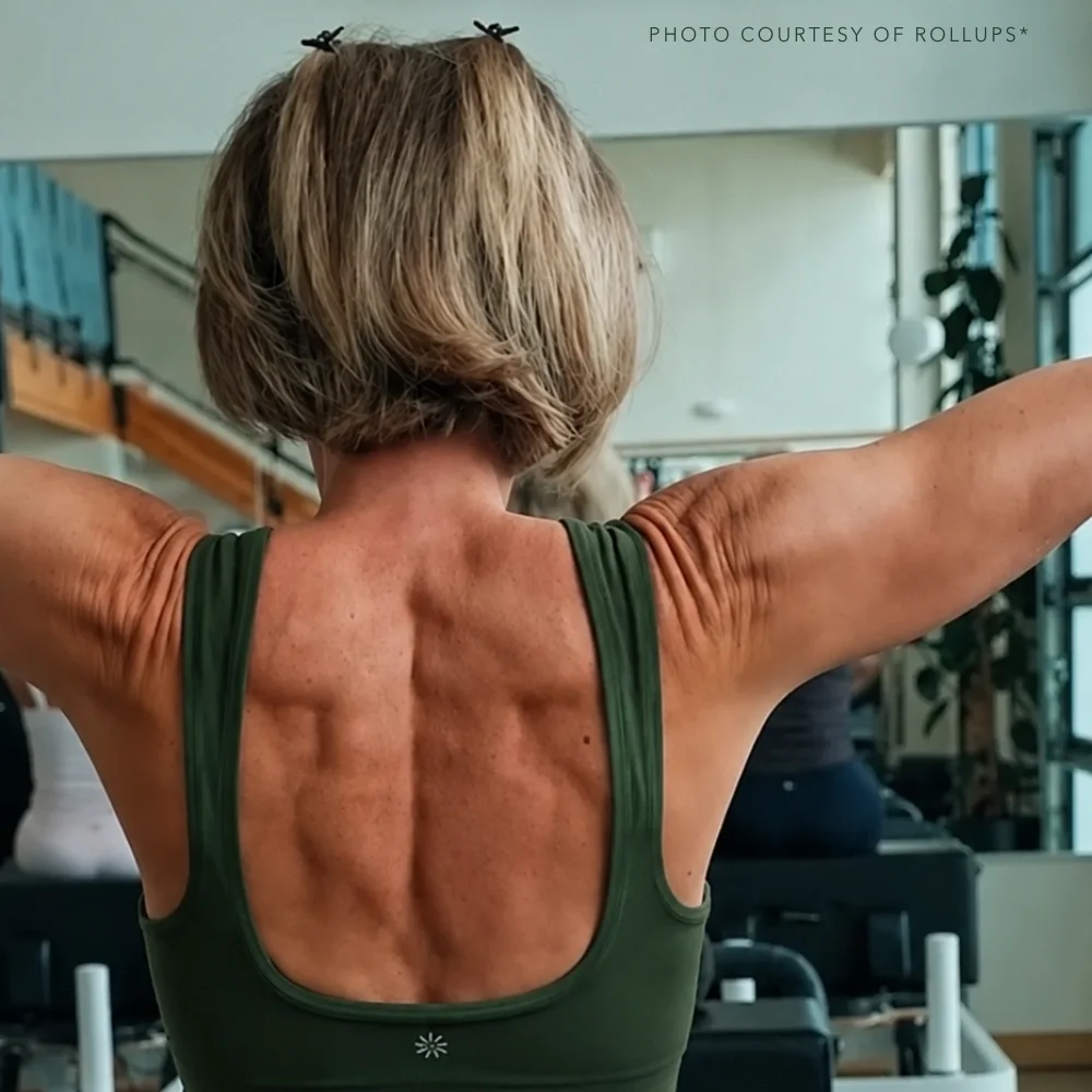 Older woman with short blonde hair stretching her arms in a gym, showing her back muscles.