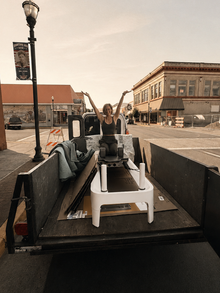 A woman sitting in a moving truck bed, exercising on a treadmill, with her arms raised, in a small town setting with buildings and street lamps.
