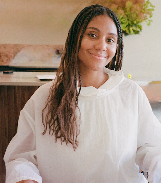 Smiling woman with long, braided hair in a white apron, standing in a kitchen with a counter and green plant in the background.