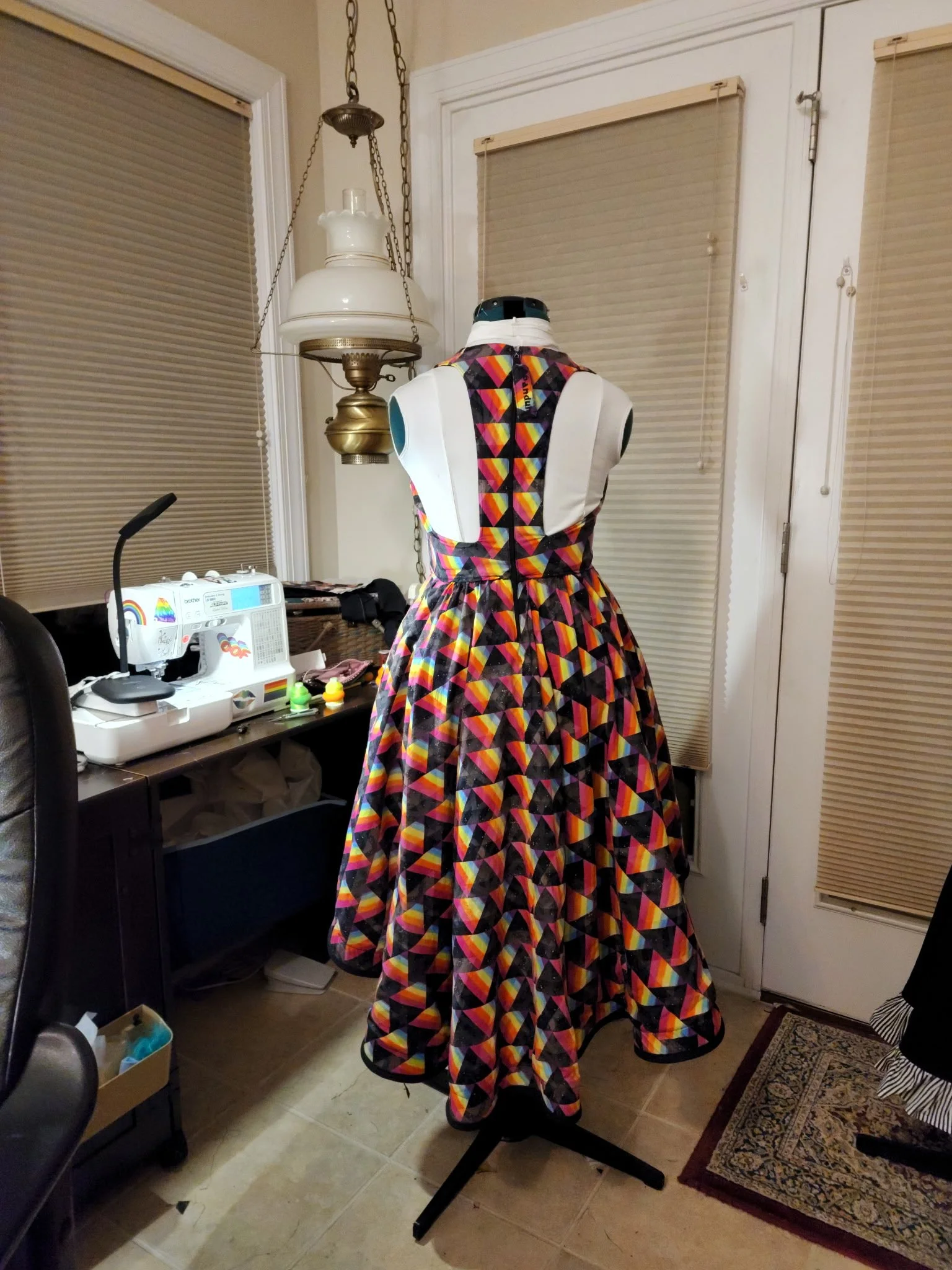 A dress displayed on a mannequin in a room adjacent to a sewing space. The dress has a geometric colorful pattern with shades of pink, orange, yellow, purple, and black. The sewing space features a Brother sewing machine with rainbow rainbow stickers, fabrics, and sewing supplies on the table.