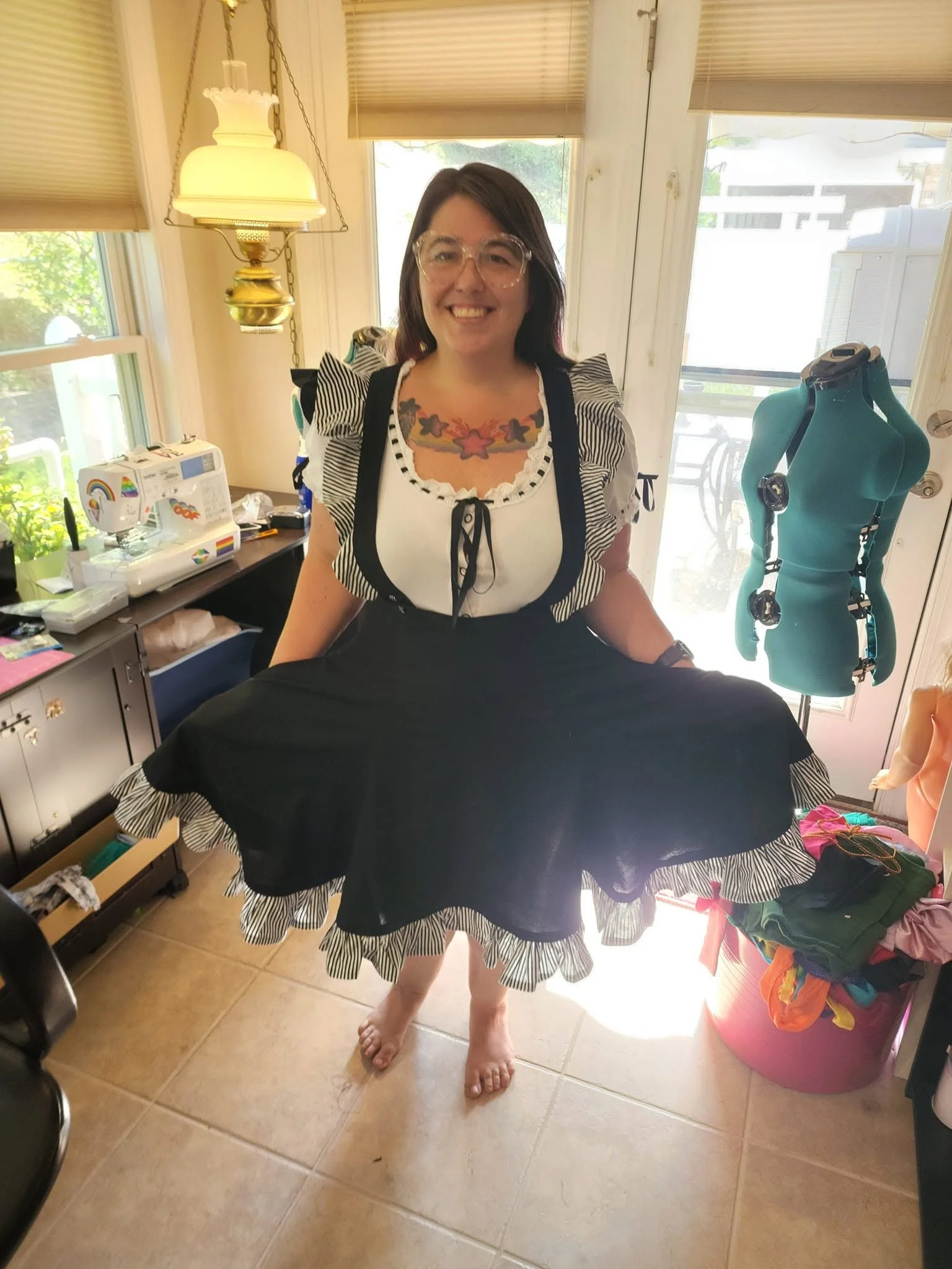 Woman standing in a brightly lit room, smiling, wearing glasses, with colorful tattoos on her chest. She is dressed in a black and white dress with ruffled sleeves and holding a large piece of black fabric. The room contains a dress form, a table with sewing supplies, and windows with beige blinds.