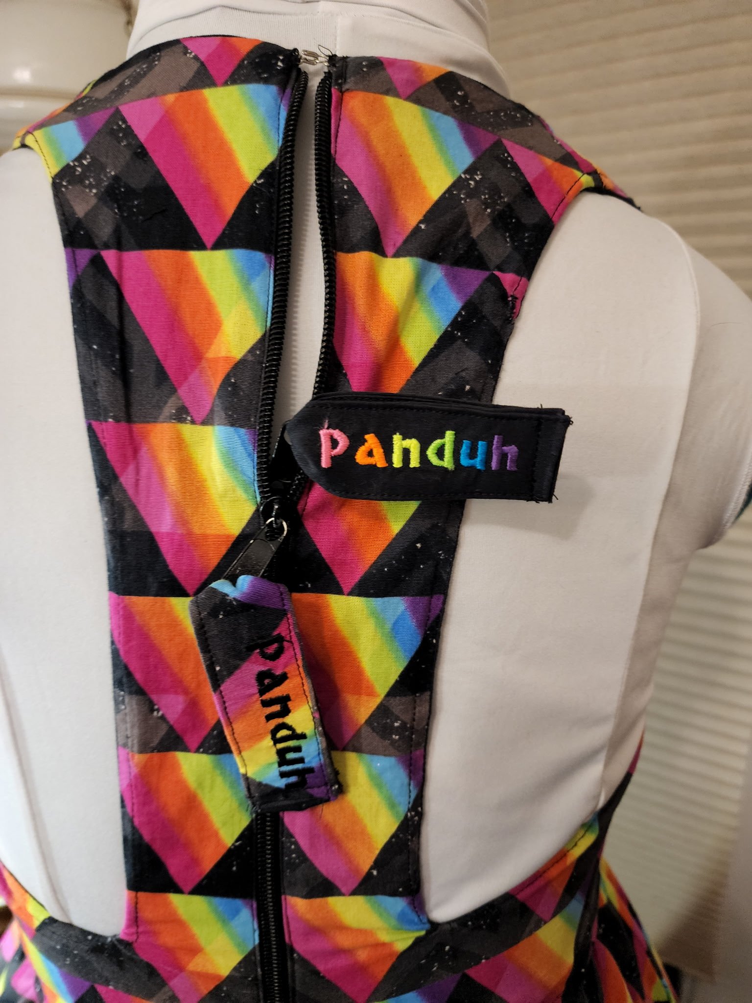 Close-up of a vest with rainbow-colored geometric patterns and a black zipper. The vest has a black patch with the name 'Pahduh' embroidered in rainbow-colored letters.
