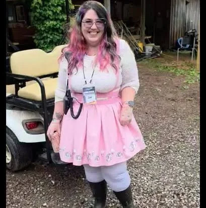 A woman with pink ombre hair, wearing glasses, a white top, pink skirt with embroidery, gray leggings, and black boots, standing outdoors near a golf cart and wooden shed.