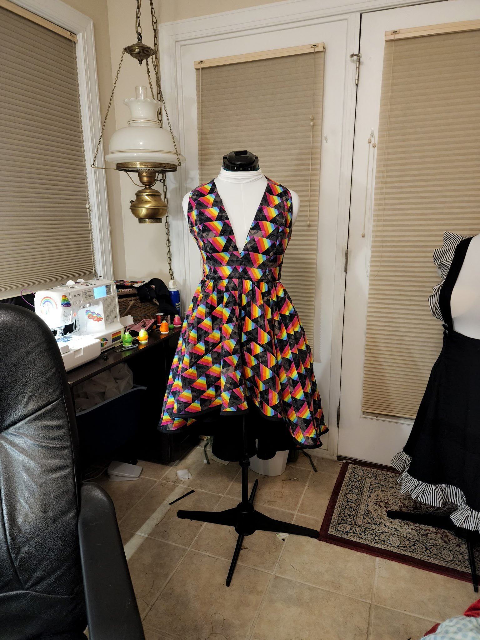 A dressmaker mannequin displaying a colorful geometric patterned dress in a room with beige tile floor, beige walls, and closed blinds over windows. To the left, there is a black desk with a sewing machine and various sewing supplies. An antique hanging lamp is above the mannequin, and a chair partially visible on the left.