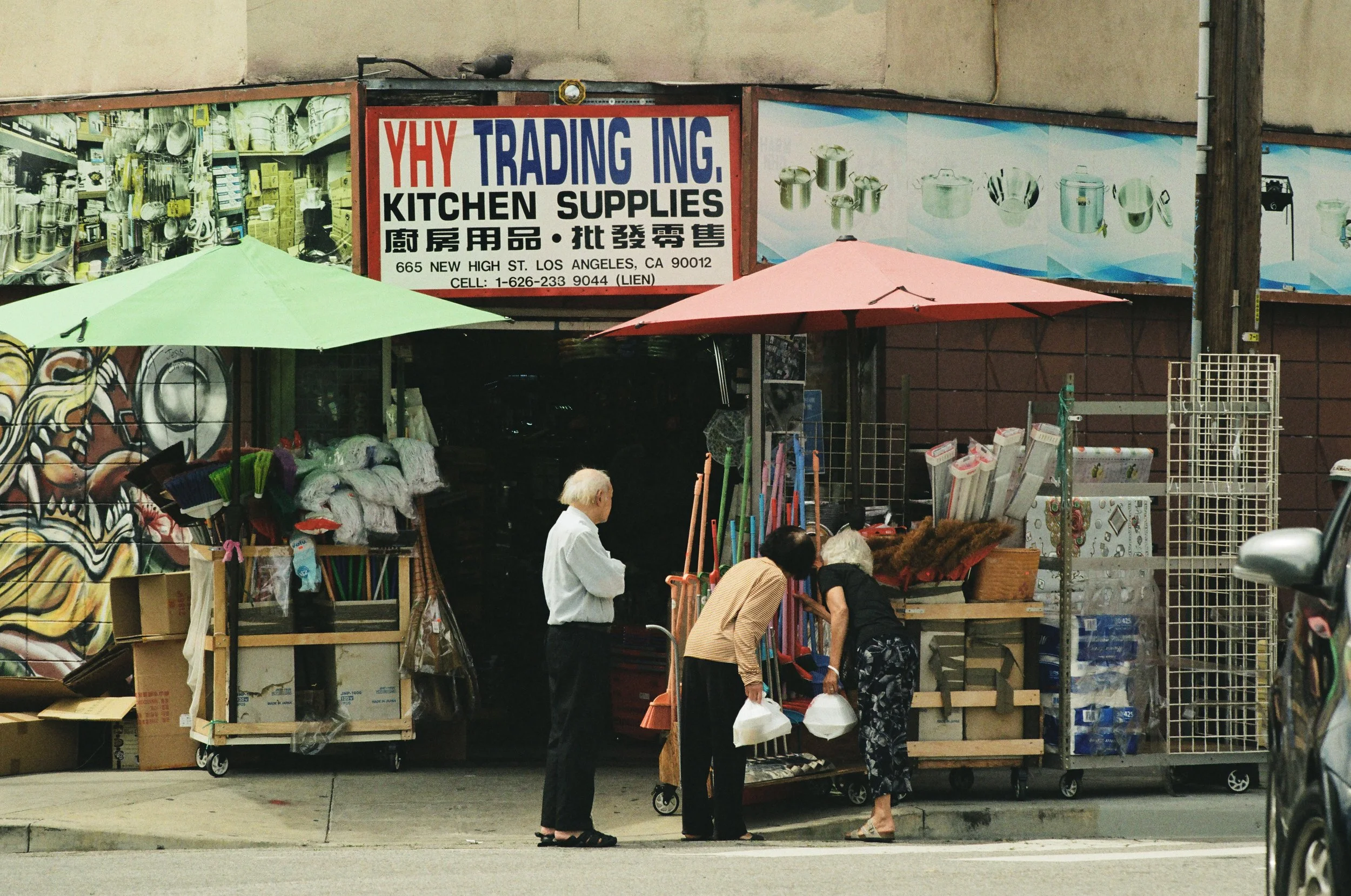 A street market stall selling kitchen supplies, including pots, brooms, and household items, with several people browsing and shopping.