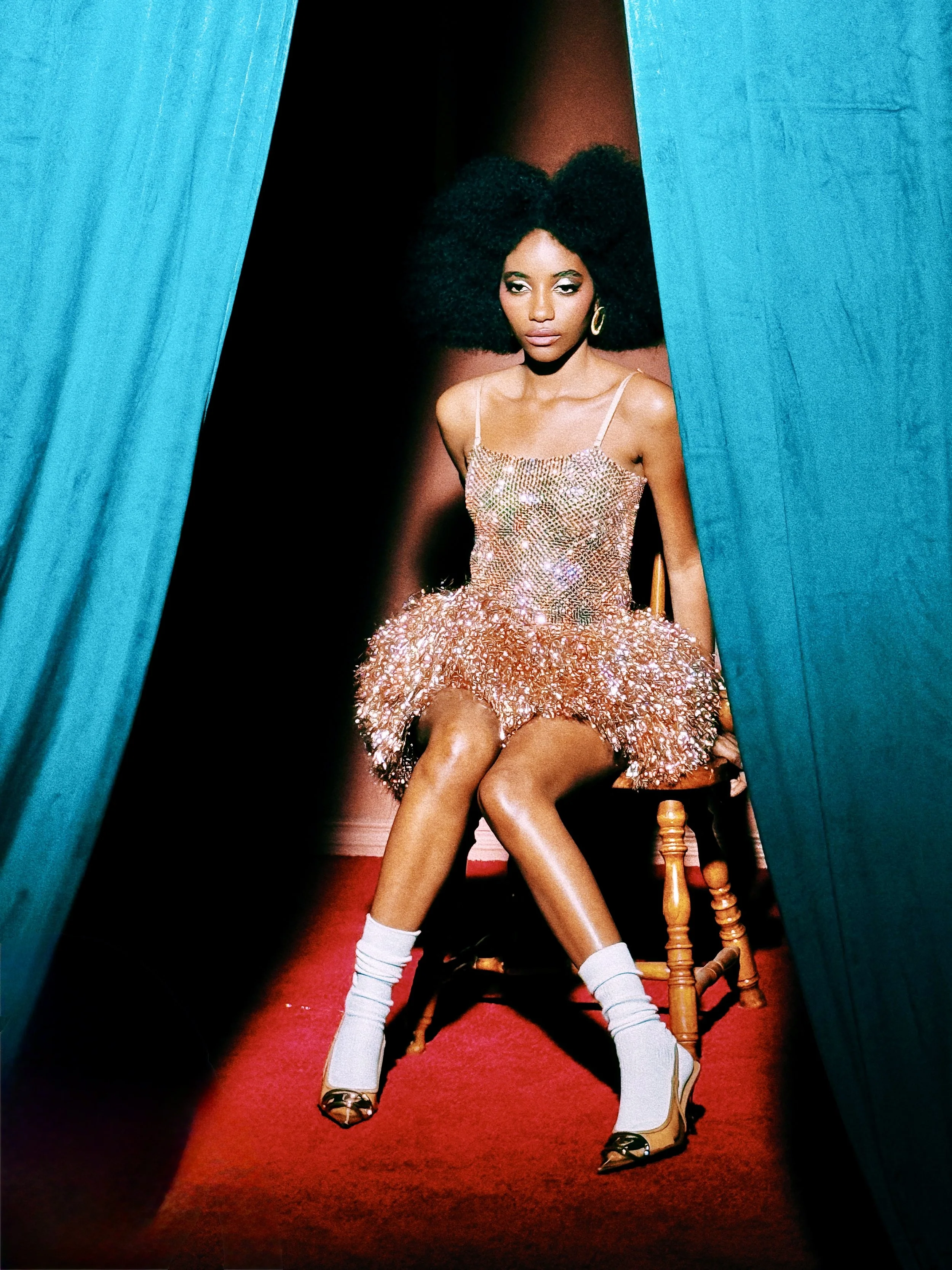 A woman with large curly hair sitting on a wooden chair, peering through a gap between blue curtains. She is dressed in a sparkly, gold dress, white knee-high socks, and high heels. A red carpet runs under her feet.