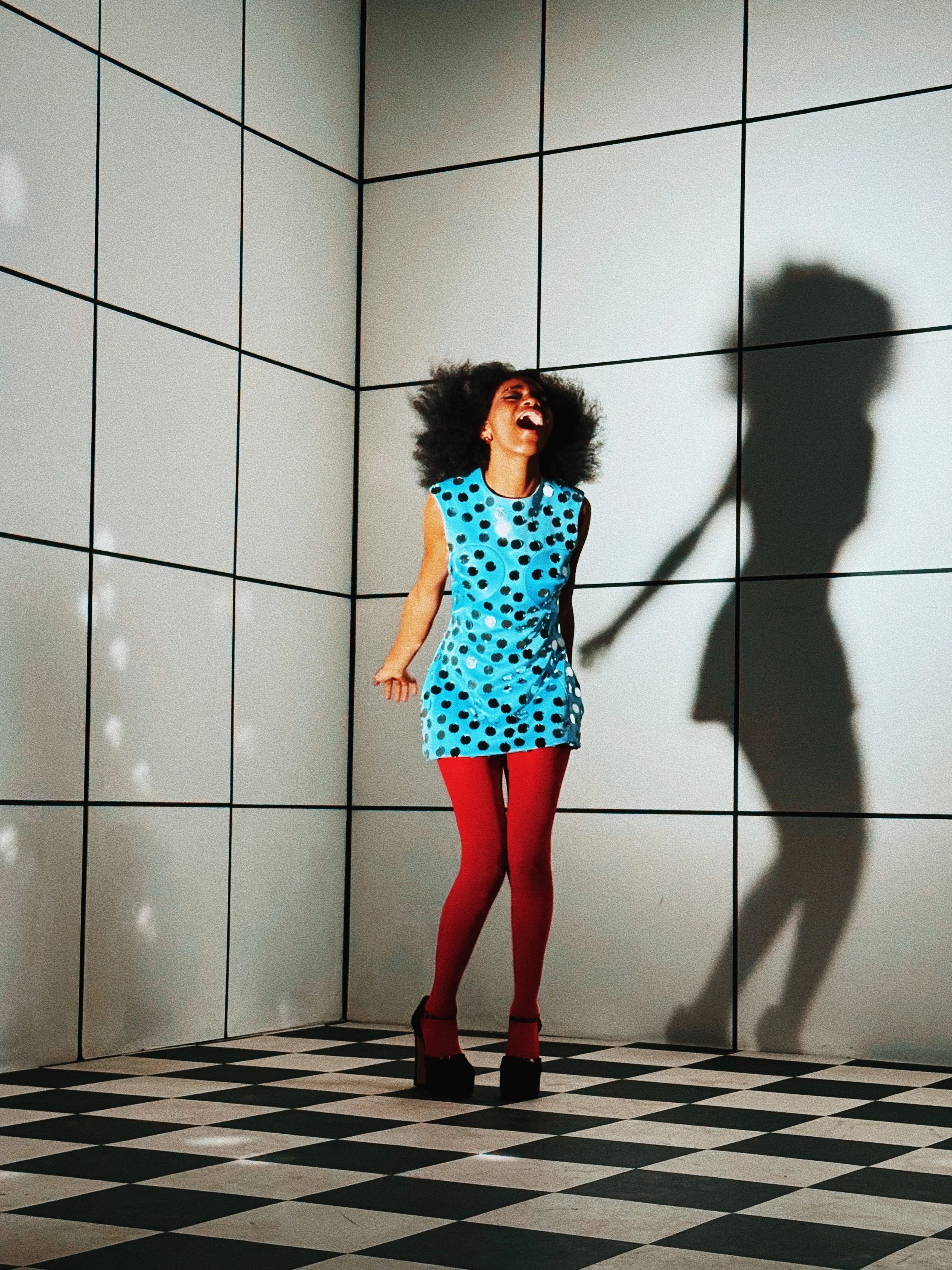 A woman with an afro hairstyle is laughing and holding her head back in a room with white tiled walls and a black-and-white checkered floor. She is dressed in a blue dress with black polka dots, red tights, and black high-heeled shoes.