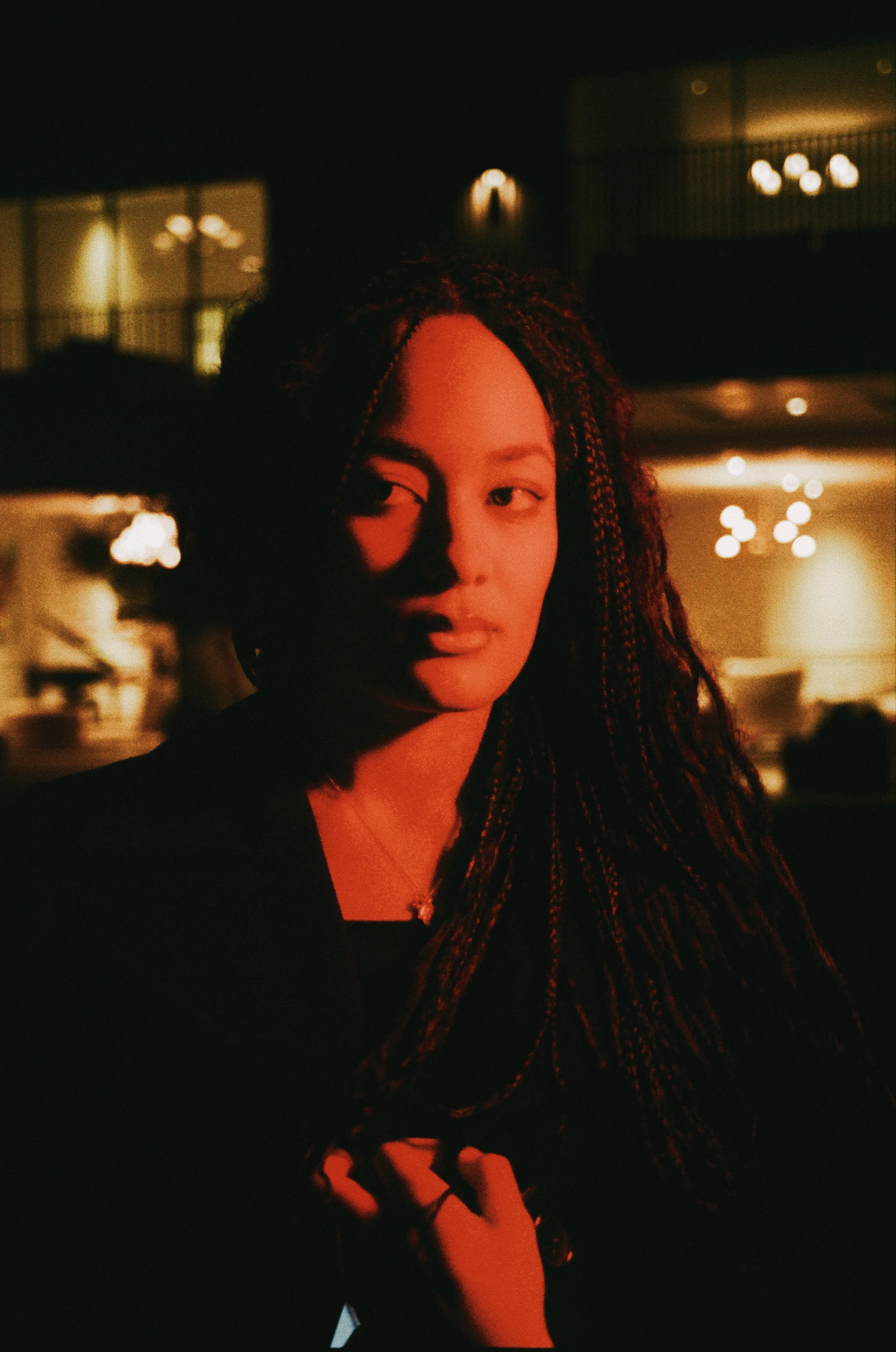 A young woman with braided hair stands in low lighting with warm tones, looking to the side. The background is blurry and dark, with some circular light reflections.