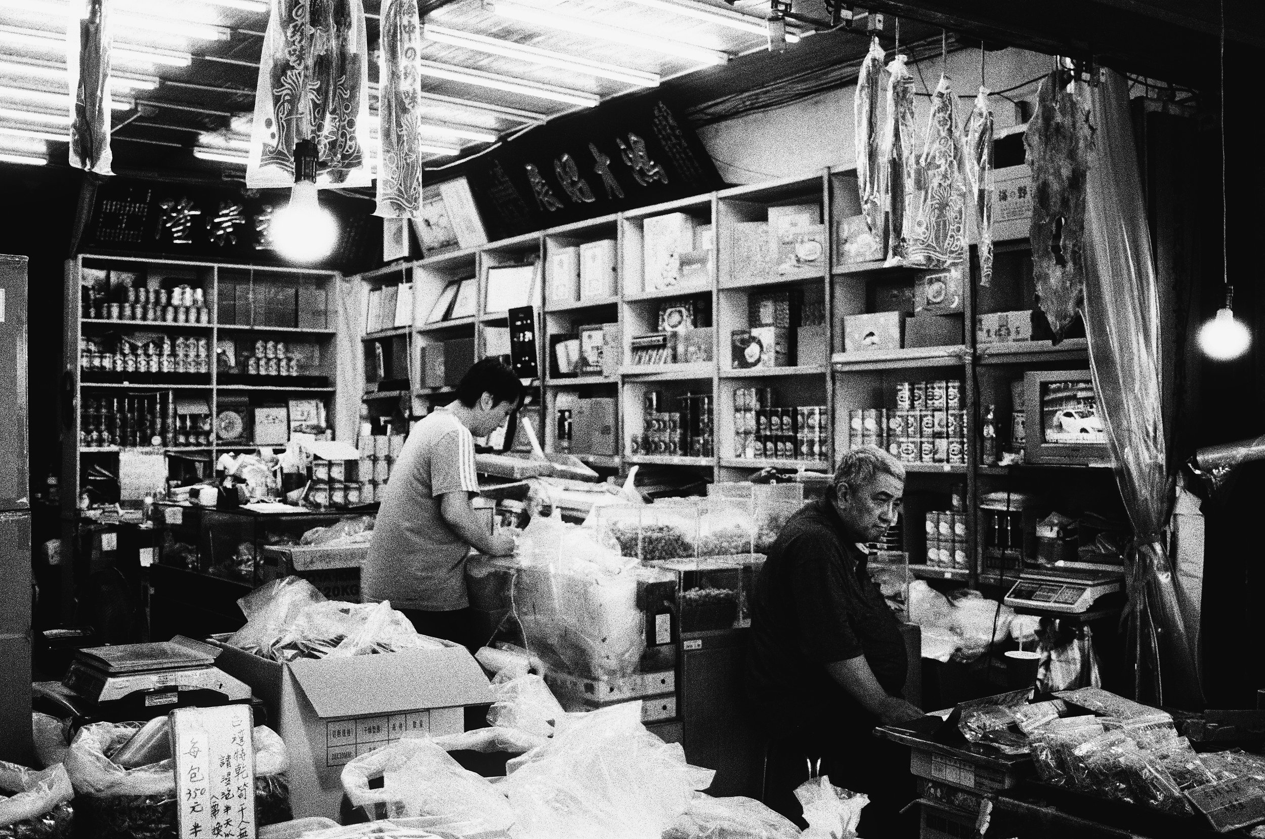 Inside a store with shelves filled with various products, two men are working at a counter, packing items or handling merchandise, while hanging fabrics or curtains decorate the ceiling.