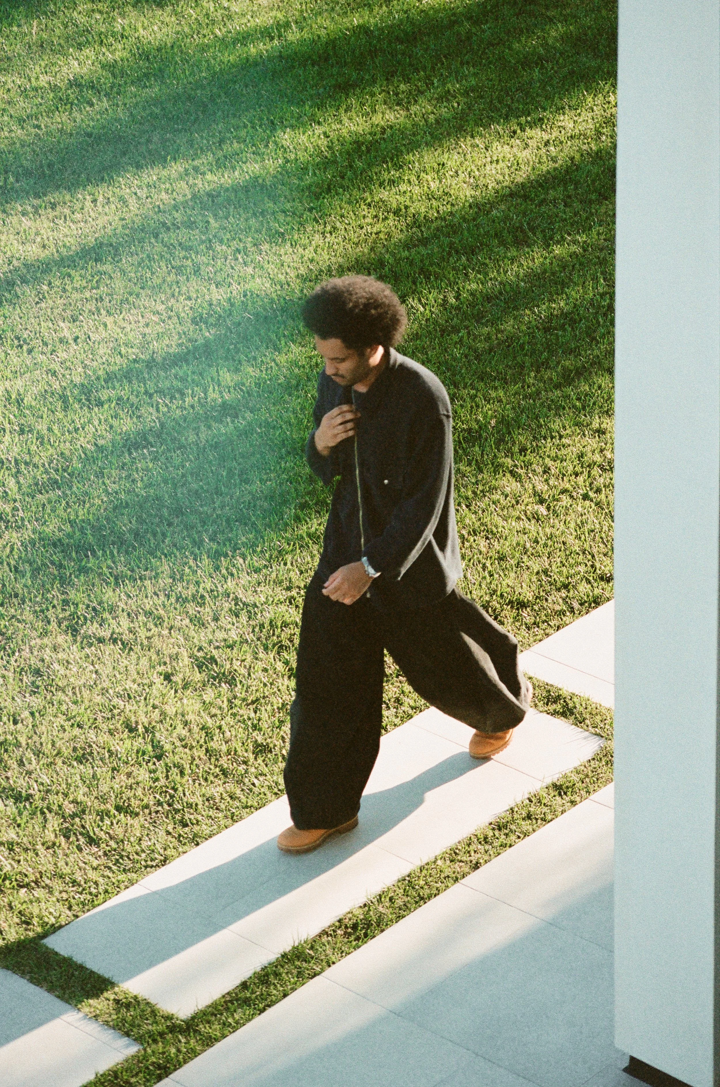 A person with curly hair walking down white steps with grass on the side, wearing a black jacket, black pants, and tan shoes.