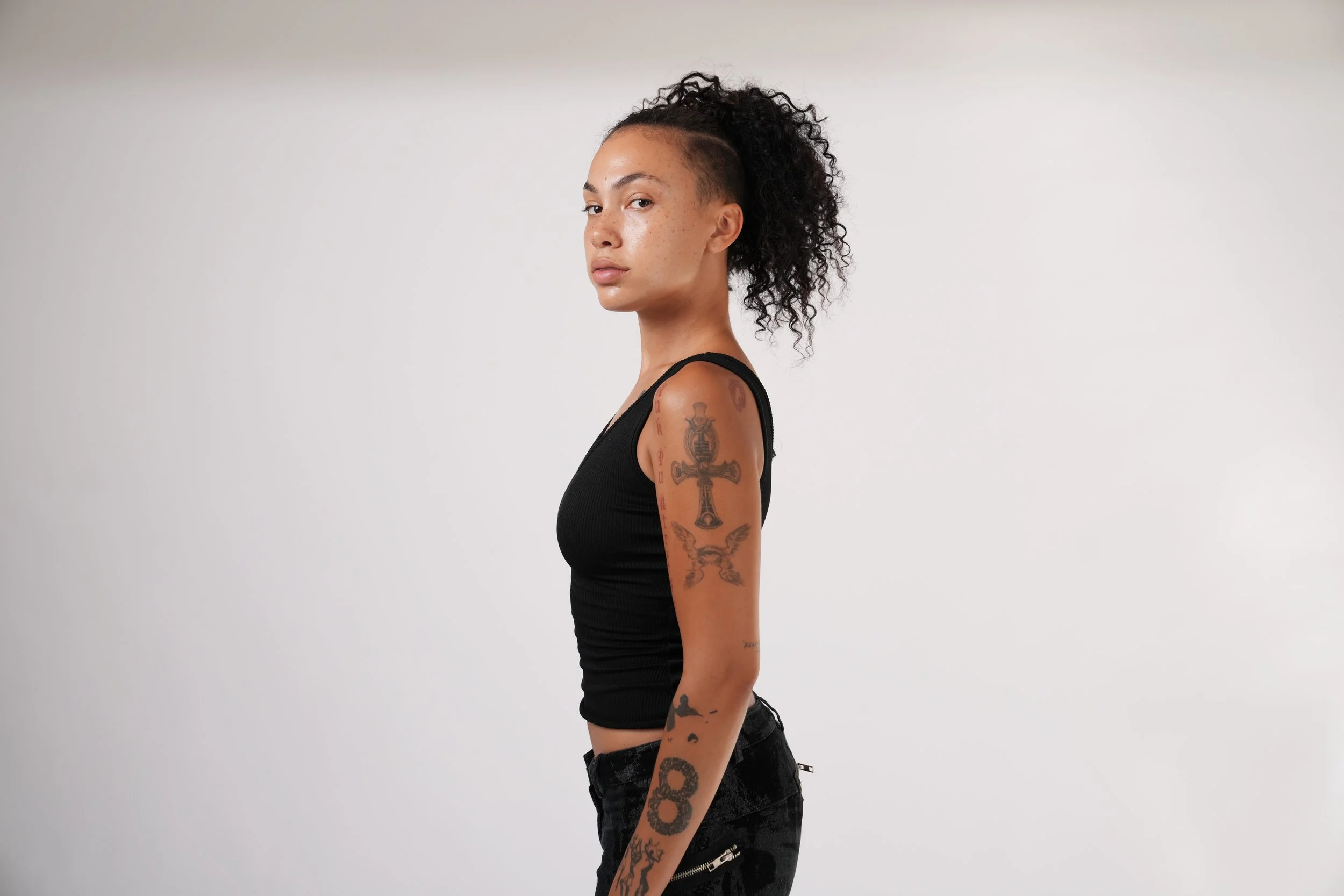 A young woman with curly dark hair and tattoos on her arm, wearing a black sleeveless top and black pants, standing sideways against a plain white background.