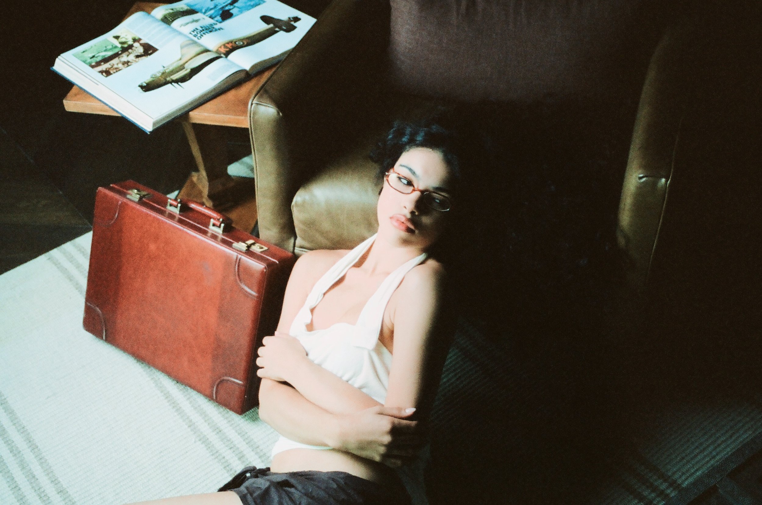 A woman with glasses lying on a green armchair in a dimly lit room. She is wearing a white top and holding a red briefcase on her lap. There is an open magazine on a wooden table beside her.