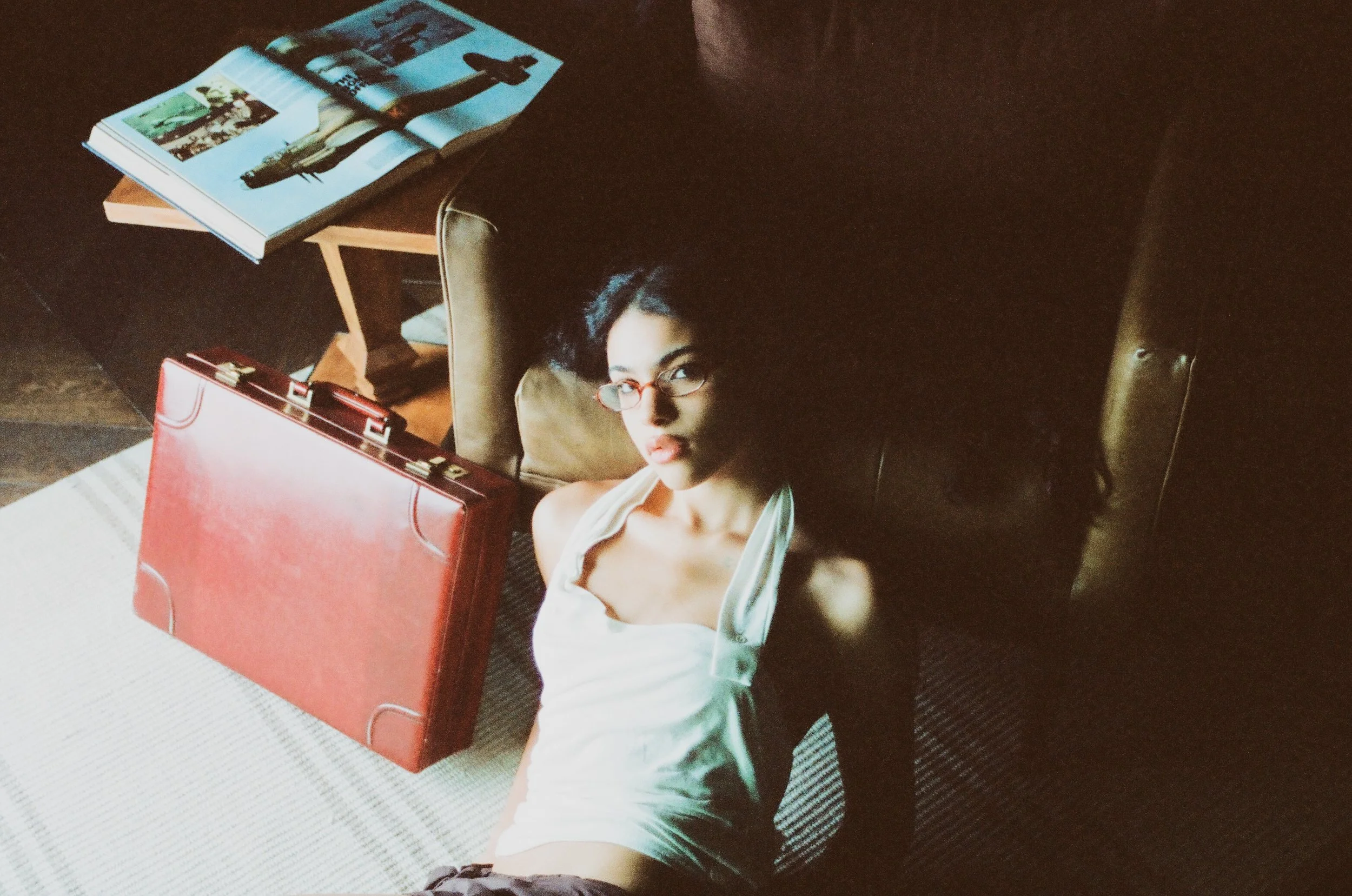 A woman with glasses lying on the floor next to a brown leather chair. There is an open magazine and a red briefcase on the floor. The woman is looking up at the camera.
