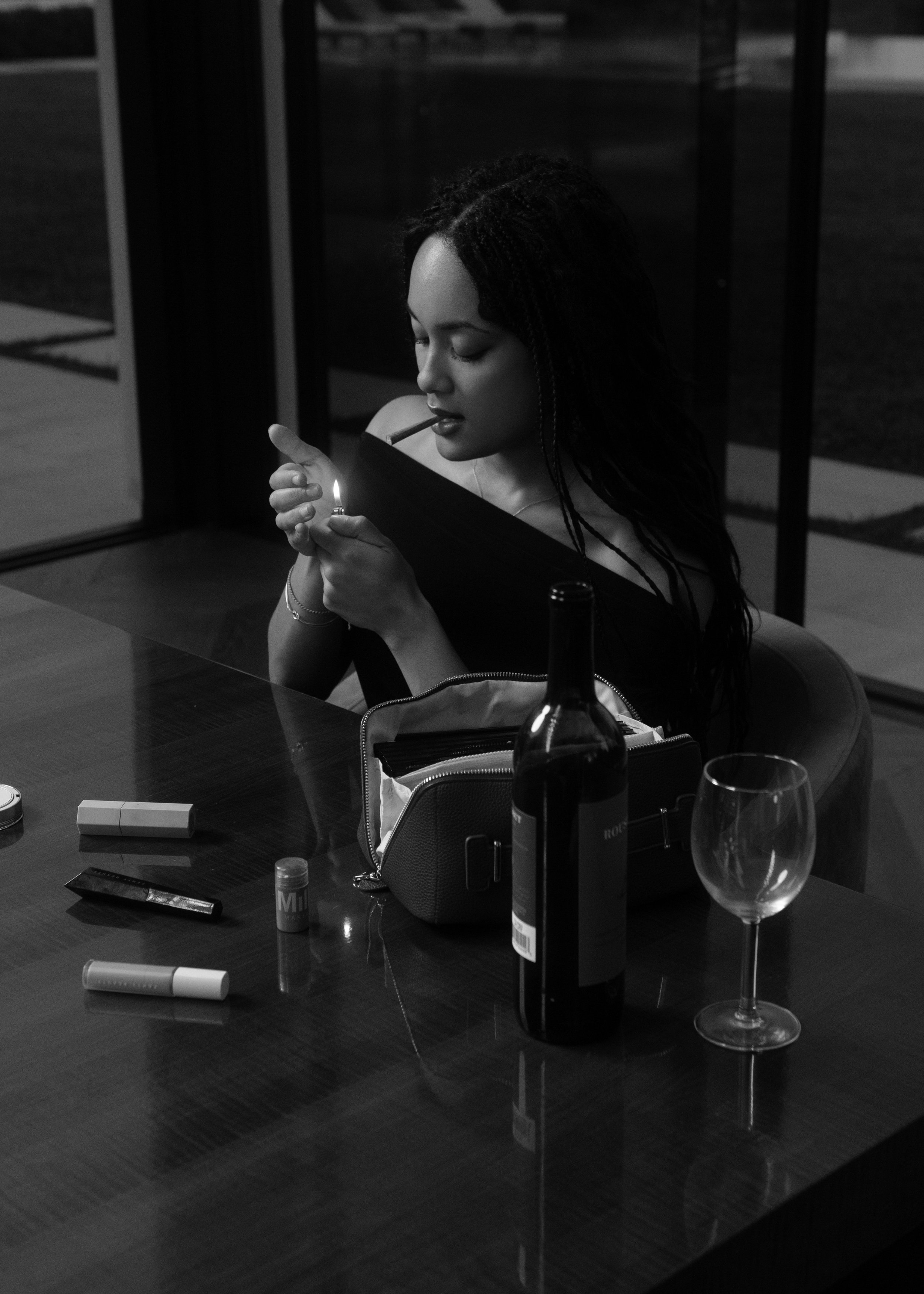Black and white photo of a woman with long, dark, braided hair seated at a table, looking at a lit cigarette in her right hand, with a lighter in her left hand. The woman has her eyes closed and appears to be in a contemplative mood. On the table in front of her are various items including a lipstick, a tube of lip gloss, a small pouch, a wine bottle, a glass with some liquid, and an empty wine glass. The background shows a glass door or window and some chairs around the table.