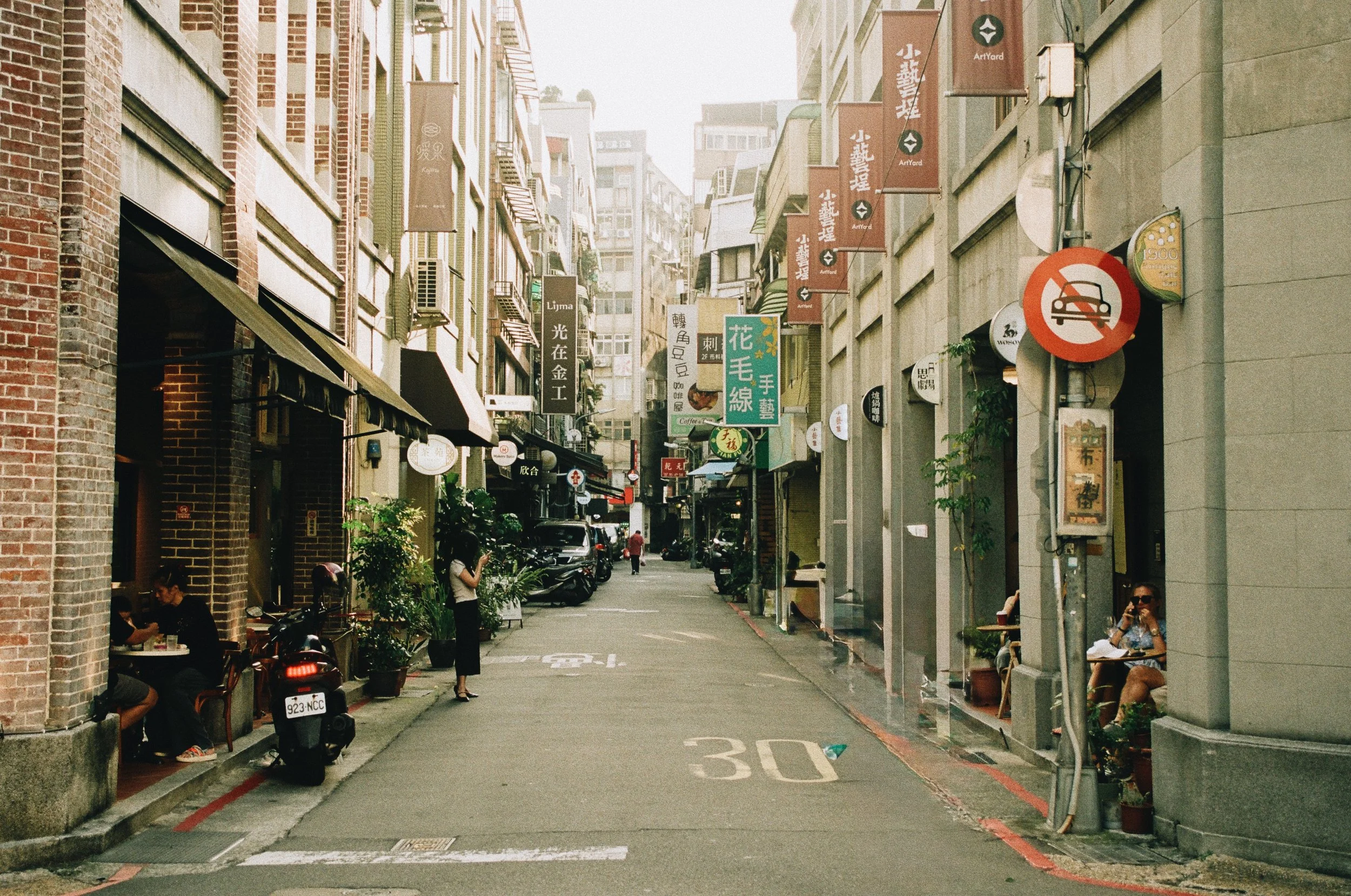 A narrow city street with parked scooters along the sidewalk, several pedestrians, and multistory buildings with various signs and banners in an urban setting.