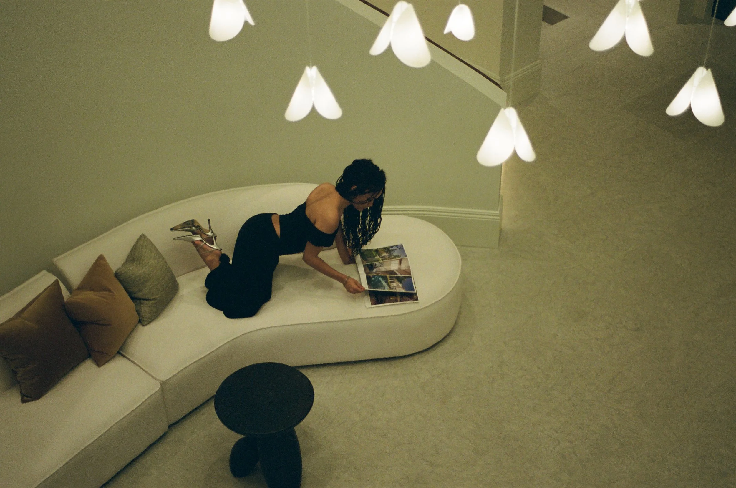 A woman with curly hair wearing a black dress and high heels is kneeling on a white couch, looking at a magazine. The couch has multiple pillows. Above, there is a modern white chandelier with multiple pendant lights hanging from the ceiling. The room has beige walls and a light-colored carpet.