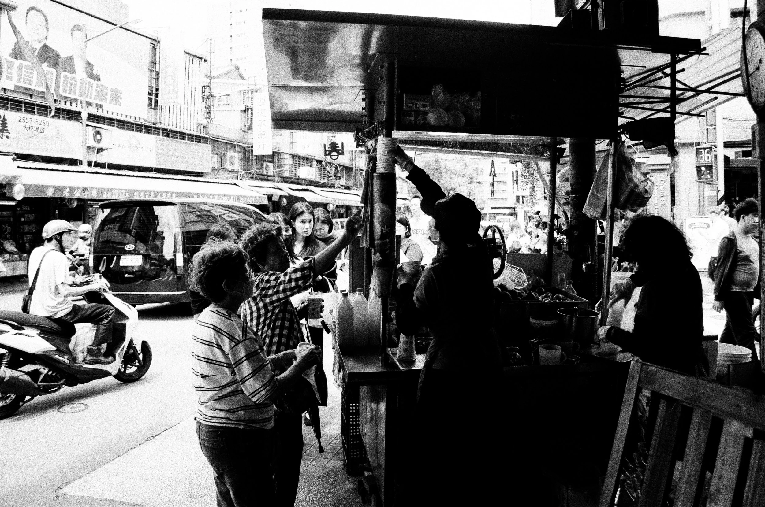 A black and white photo of a busy street market with people shopping and vendor stalls, including a person handing something across a counter, with buildings and advertisements in the background.