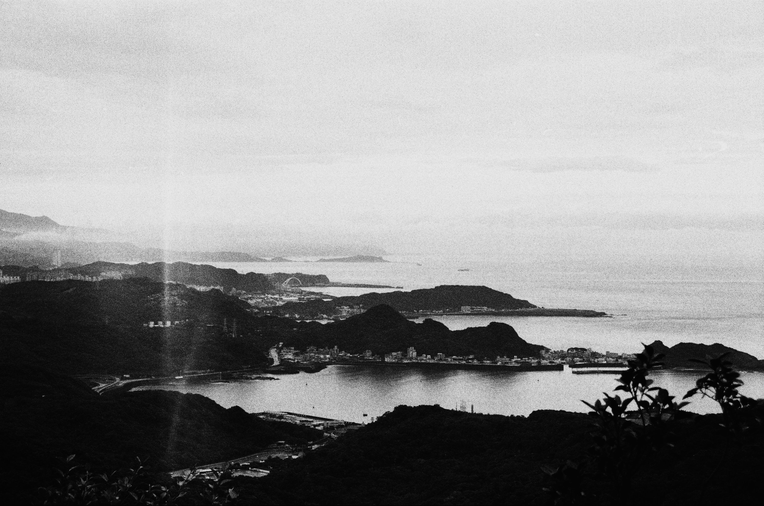 A black and white landscape photo of mountains, water, and a distant city with a Ferris wheel.