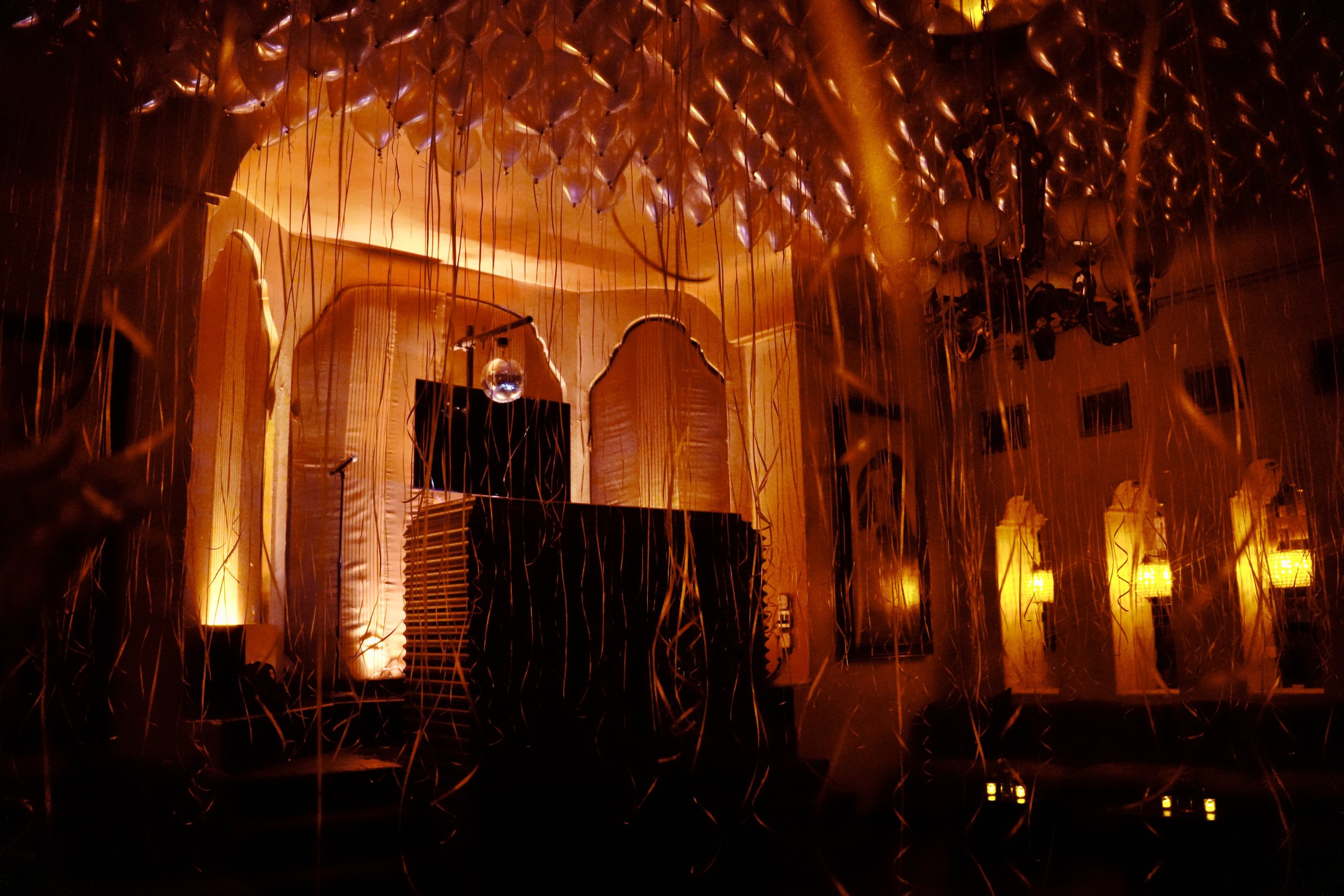 A dimly lit indoor stage with a DJ setup, including a laptop and speakers, surrounded by numerous rose gold balloons and hanging string decorations. Warm orange lighting illuminates the scene.
