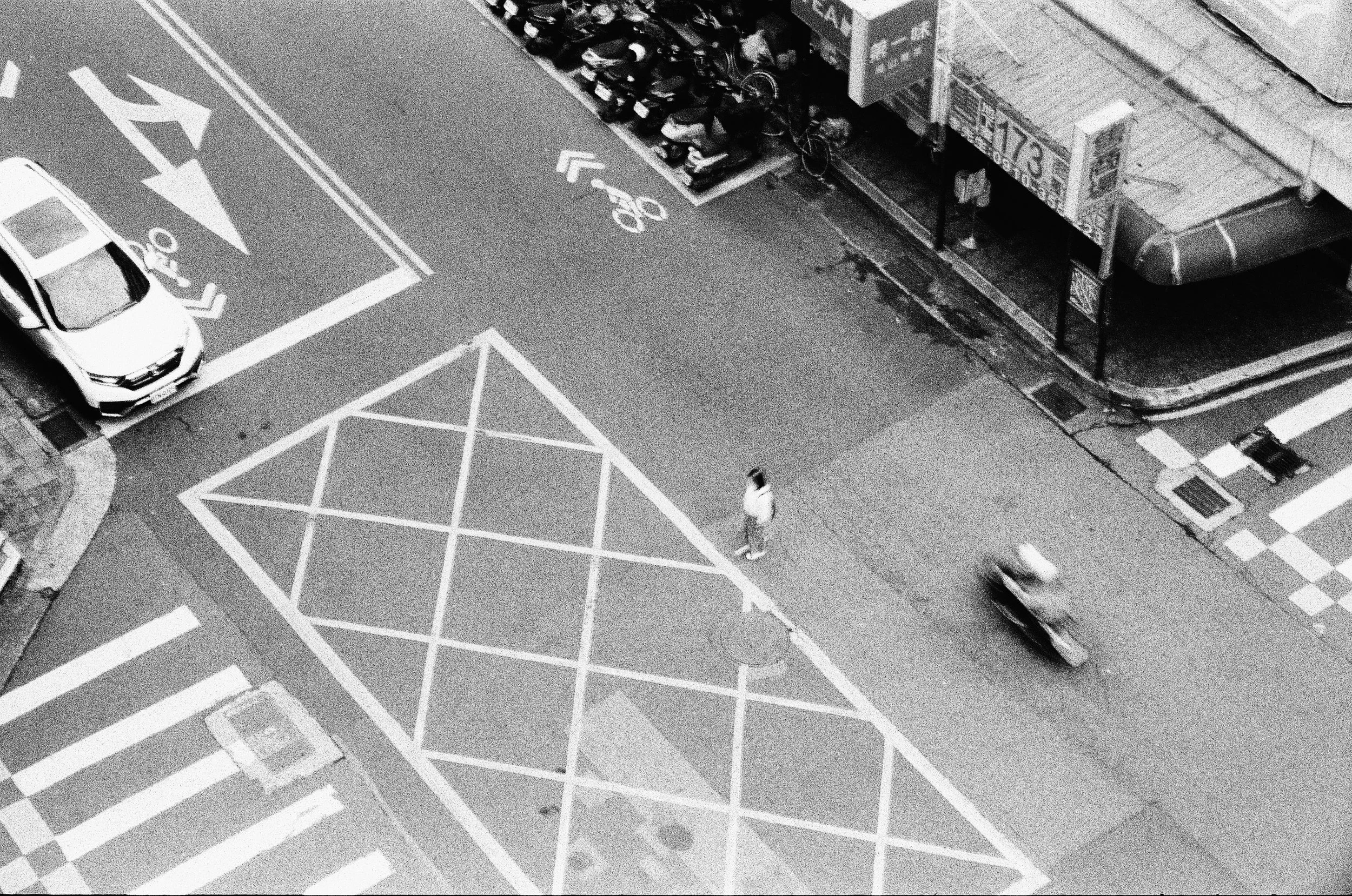A black and white photo of a city street intersection with marked lanes, a parked car, and a person walking near the sidewalk. There are bikes parked against a building and a truck nearby.