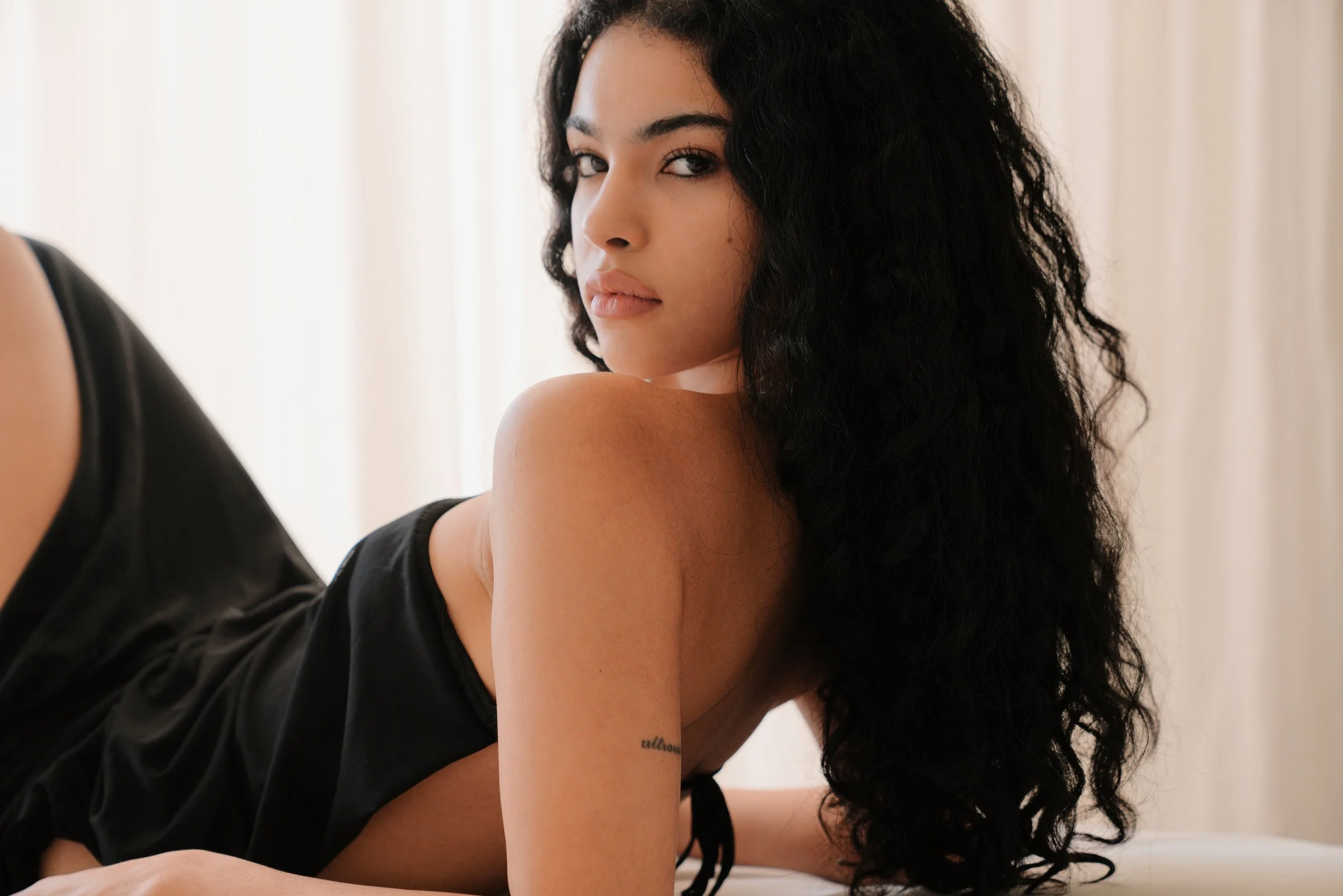 A woman with long, curly black hair and light skin looking over her shoulder at the camera, with a tattoo on her arm, wearing a black top.