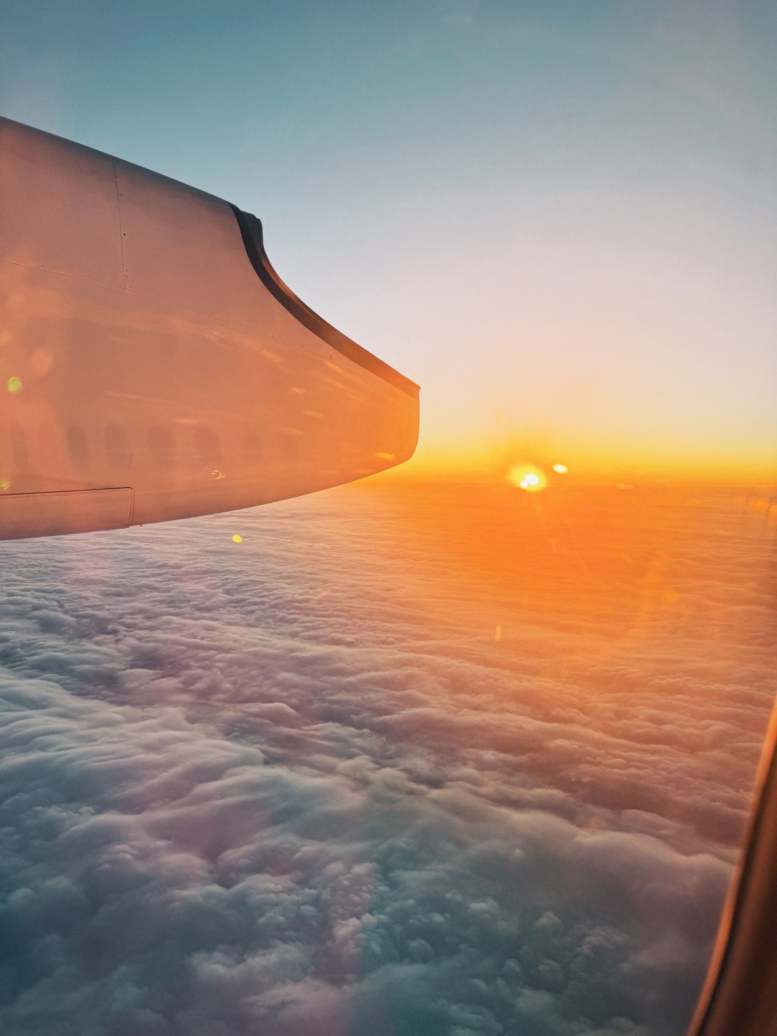 Photo taken from an airplane window showing the aircraft wing, clouds below, and a sunset or sunrise sky with the sun near the horizon.