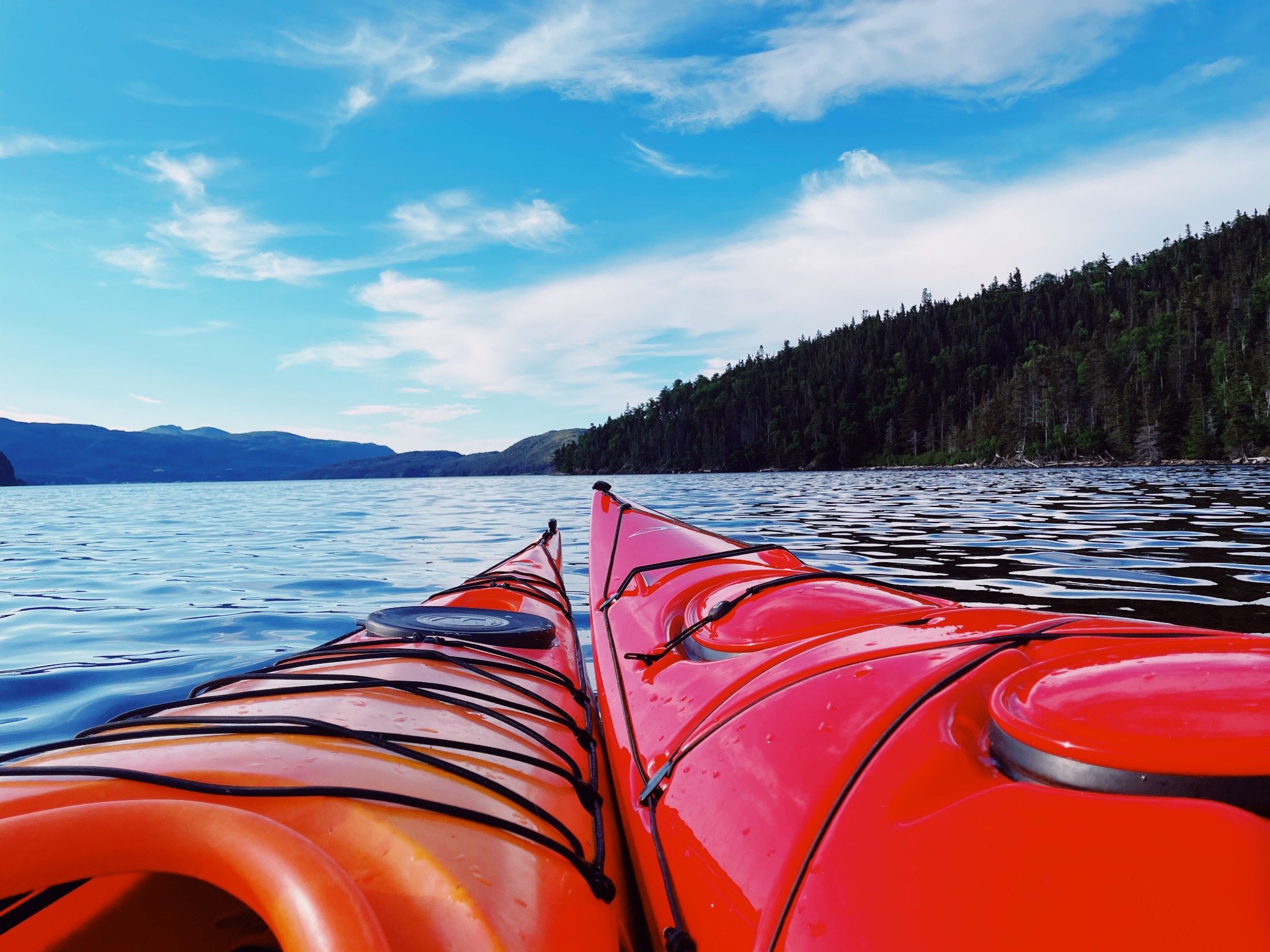 View from a red kayak on a large lake with forested hills in the background under a partly cloudy sky.