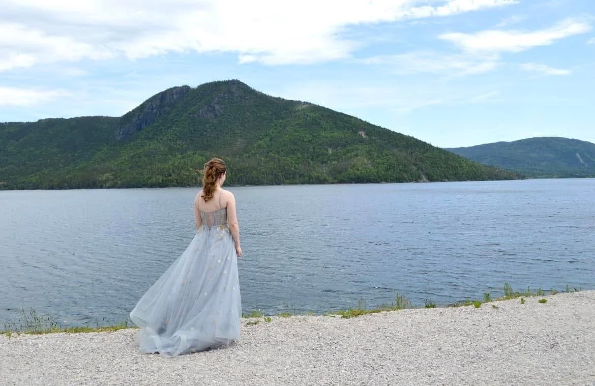 A woman in a flowing, light-colored dress standing on the gravel shore and looking out at a lake with green mountains in the distance under a partly cloudy sky.