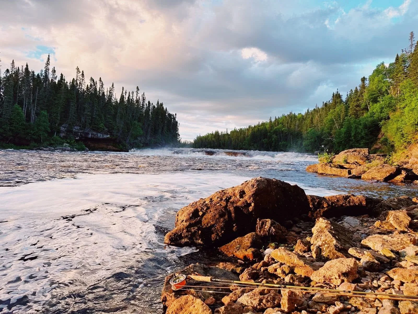 River flowing through a forested area with rocks in the foreground and a cloudy sky above.